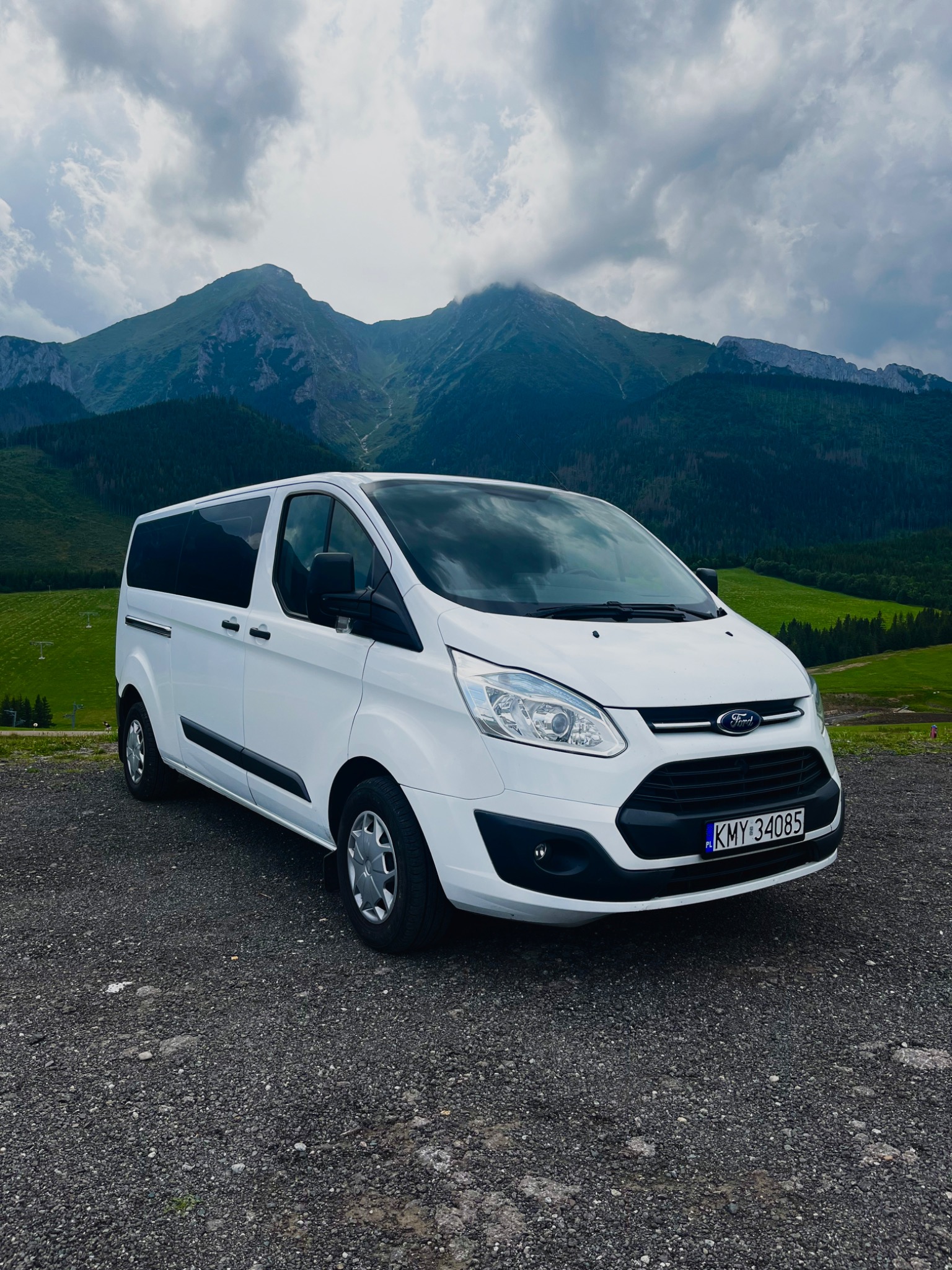 Biały bus Ford Transit na tle górskiego krajobrazu z pochmurnym niebem. Widoczne tablice rejestracyjne KMY 34085.