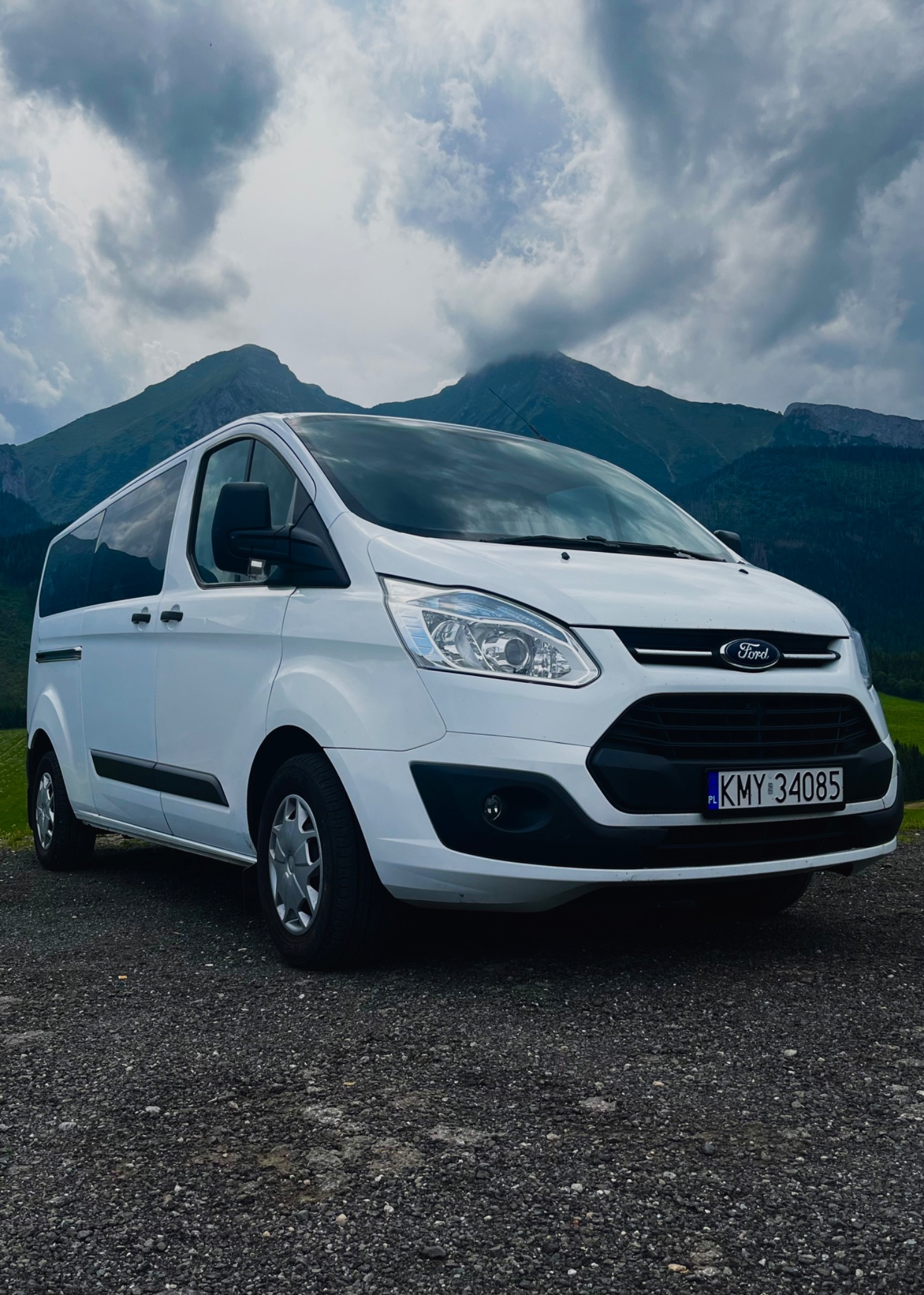Biały Ford Transit z polską tablicą rejestracyjną KMY 34085 na tle górskiego krajobrazu z pochmurnym niebem.