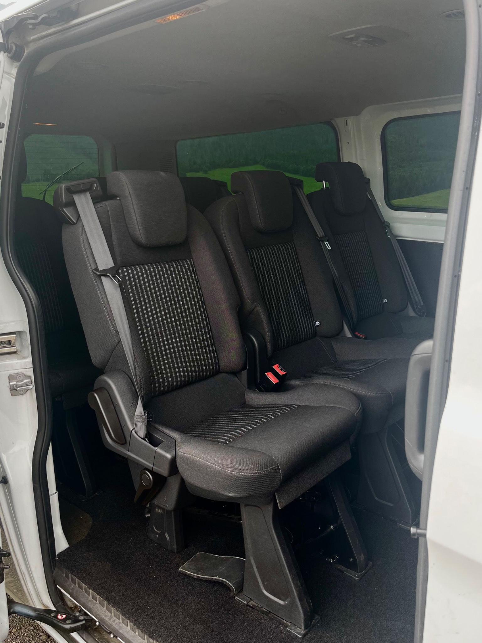 Wnętrze białego busa z trzema rzędami czarnych foteli z pasami bezpieczeństwa, widok z otwartych drzwi bocznych na zielony krajobraz za oknem.