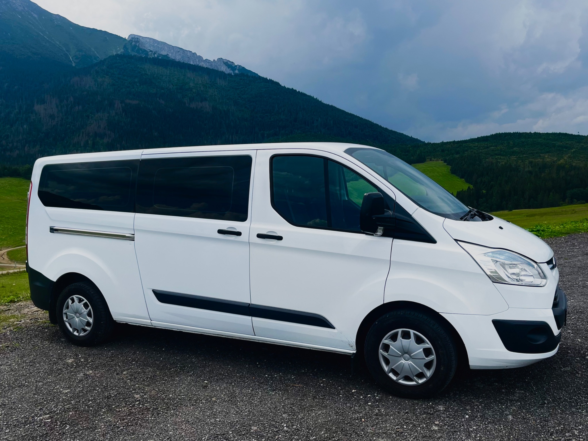 Biały bus Ford Transit na tle górskiego krajobrazu w pochmurny dzień.