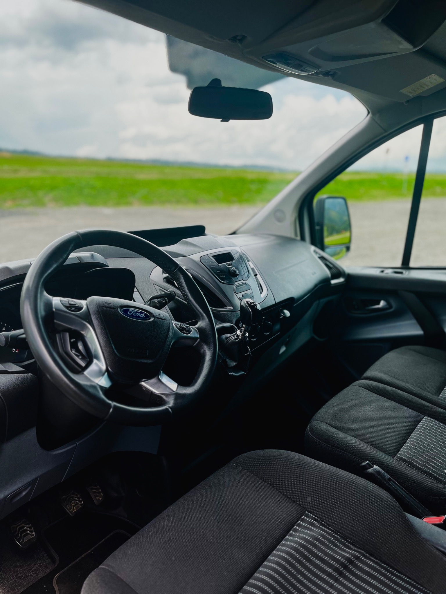 Wnętrze busa marki Ford Transit z widokiem na kierownicę, deskę rozdzielczą i przednie fotele z szarymi paskami, za szybą rozciąga się zielony krajobraz.