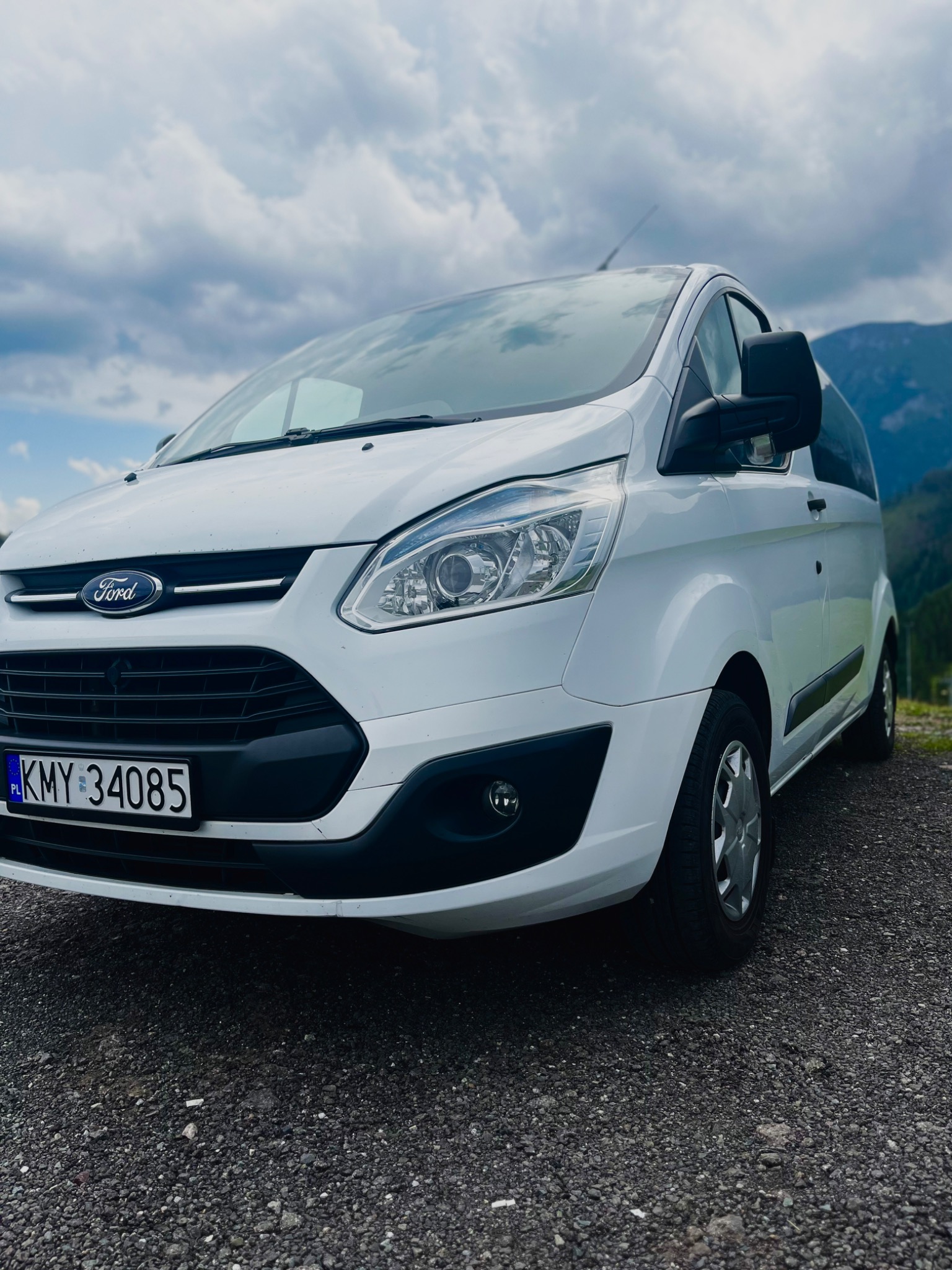 Biały Ford Transit z polską tablicą rejestracyjną KMY 34085, zaparkowany na żwirowej nawierzchni z widokiem na góry w tle.