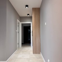 Michał Wielgus - Korytarz po remoncie: szare ściany z fakturą, beżowe płytki podłogowe, białe listwy przypodłogowe, drewniana szafa wnękowa i czarne lampy sufitowe.