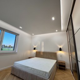 Michał Wielgus - Nowoczesna sypialnia po remoncie: jasne ściany, parkiet w jodełkę, beżowe łóżko z tapicerowanym zagłówkiem, przesuwne drzwi szafy i okno z widokiem na zieleń.