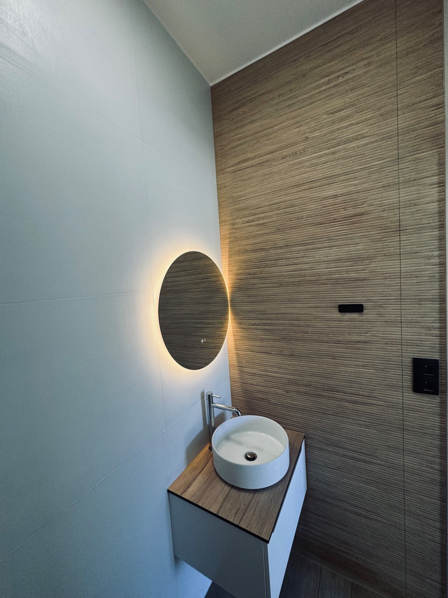 Nowoczesna łazienka z okrągłym lustrem podświetlanym LED, umywalką nablatową na szafce z drewnianym blatem i płytkami imitującymi drewno na ścianie.