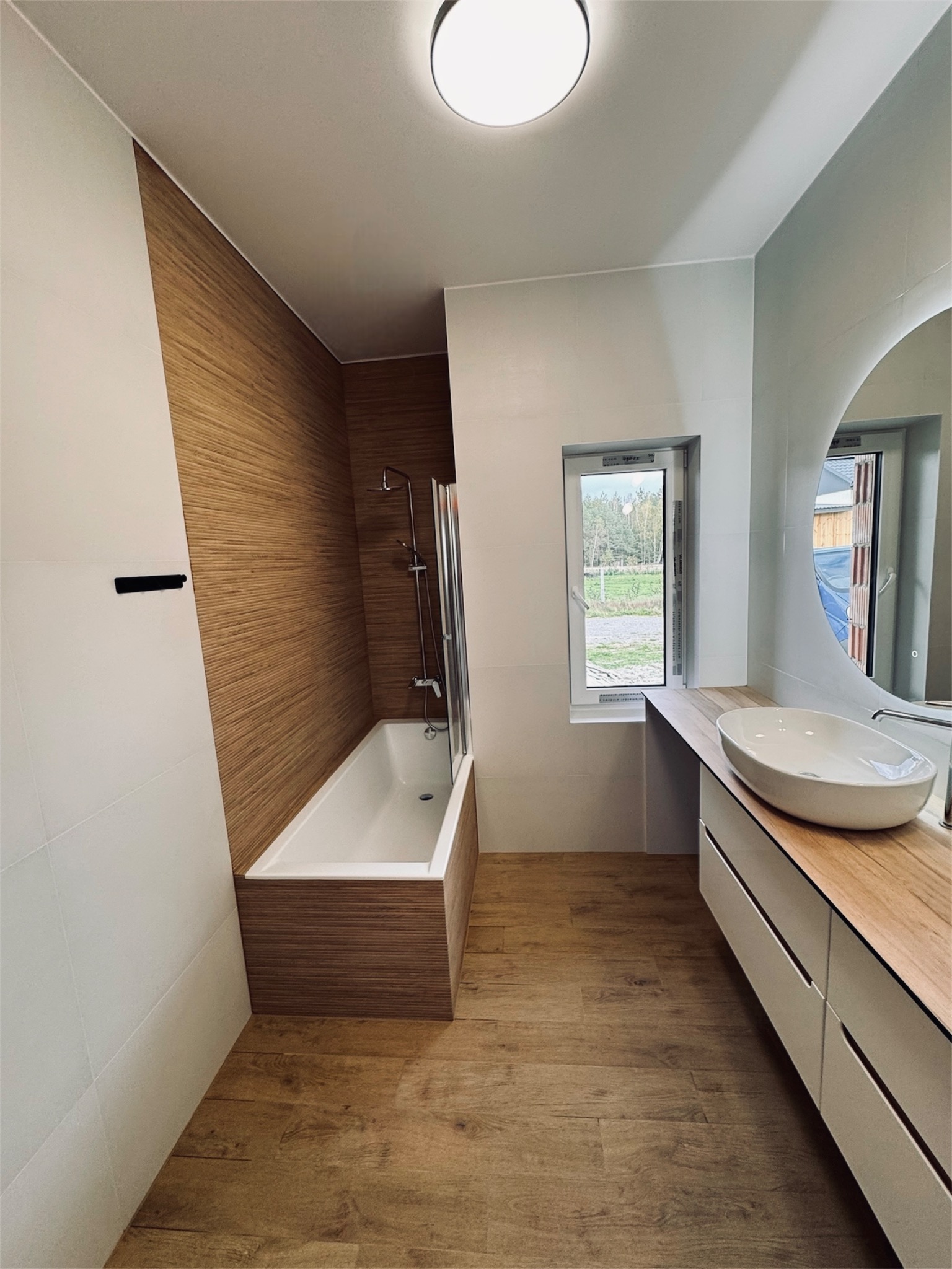 Nowoczesna łazienka z drewnianymi akcentami: panelowa ściana przy wannie, drewnopodobna podłoga, biała umywalka nablatowa i szafka z drewnianym blatem, okrągłe lustro z podświetleniem.