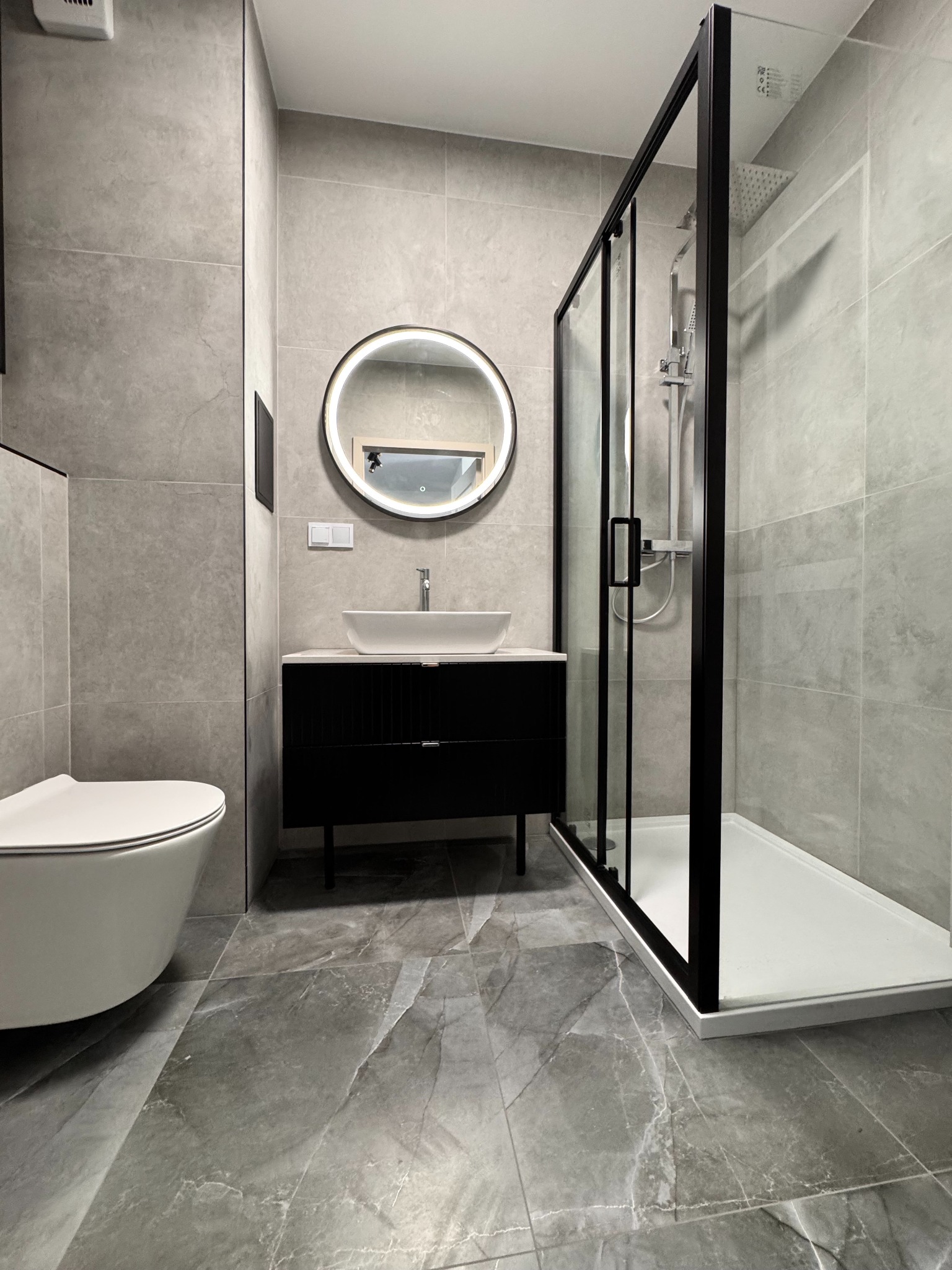 Elegancka łazienka w szarościach: kabina prysznicowa z czarnymi ramami, okrągłe lustro z podświetleniem nad białą umywalką na czarnej szafce, marmurowe płytki.
