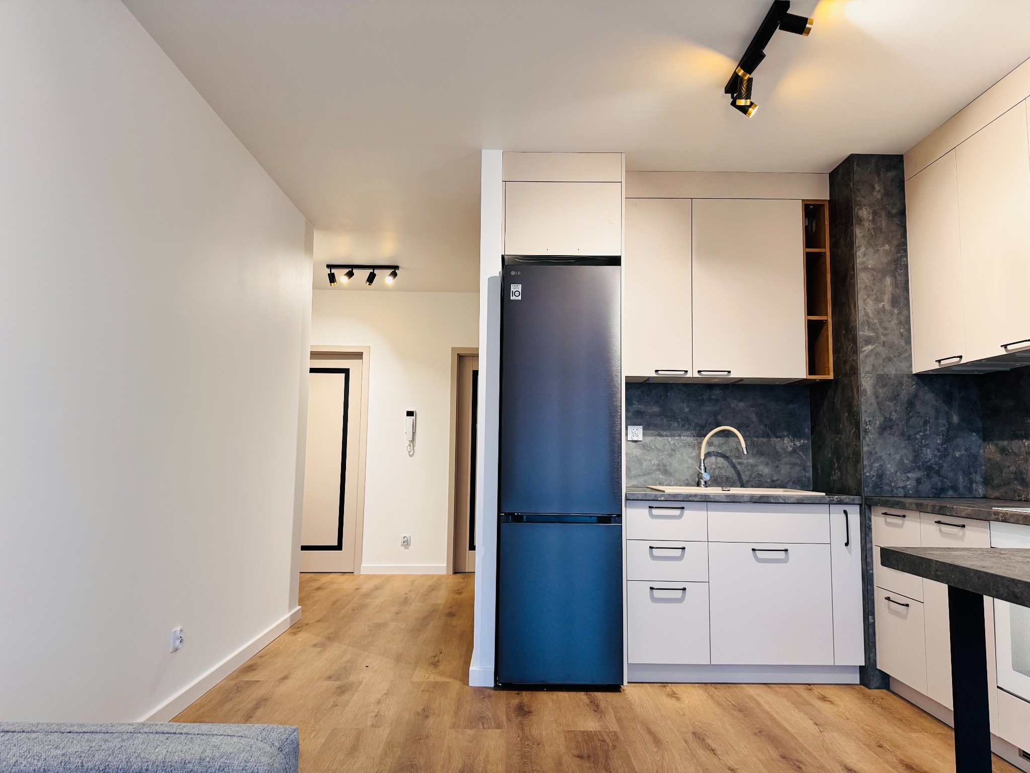 Nowoczesna, jasna kuchnia z drewnianą podłogą, białymi szafkami i grafitową lodówką LG. Oświetlenie punktowe podkreśla minimalistyczny design wnętrza. Widok na salon.