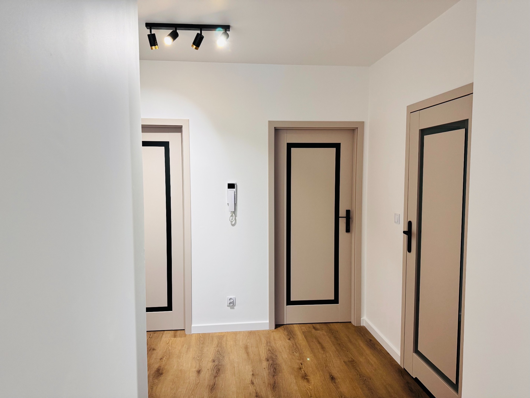 Jasne wnętrze po remoncie: drewnopodobna podłoga, białe ściany, trzy beżowe drzwi z czarnymi obramowaniami i oświetlenie punktowe.