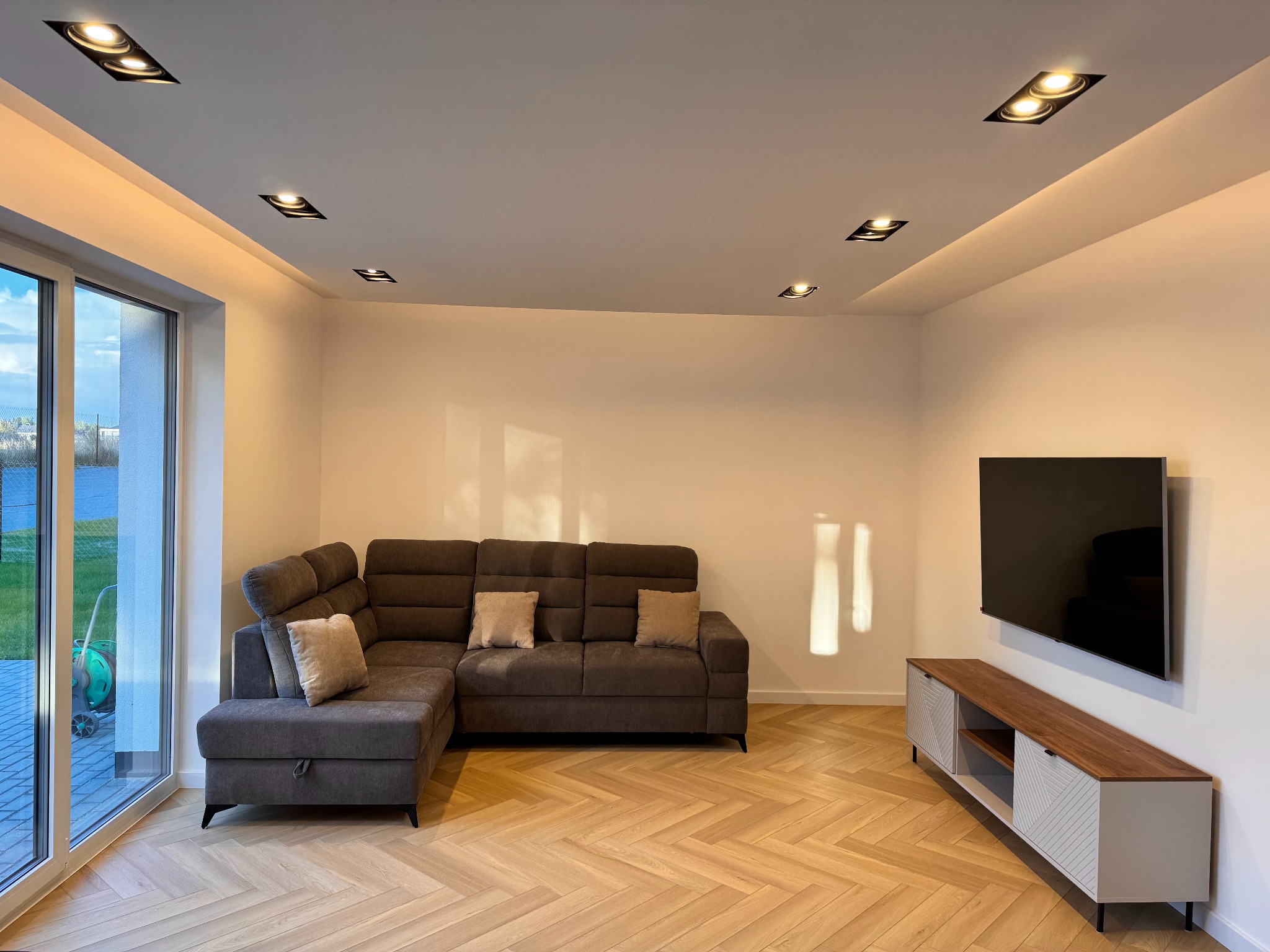 Jasny salon z parkietem w jodełkę, szarą sofą narożną i szafką RTV. Widoczne punktowe oświetlenie sufitowe i duże okno z widokiem na zewnątrz.