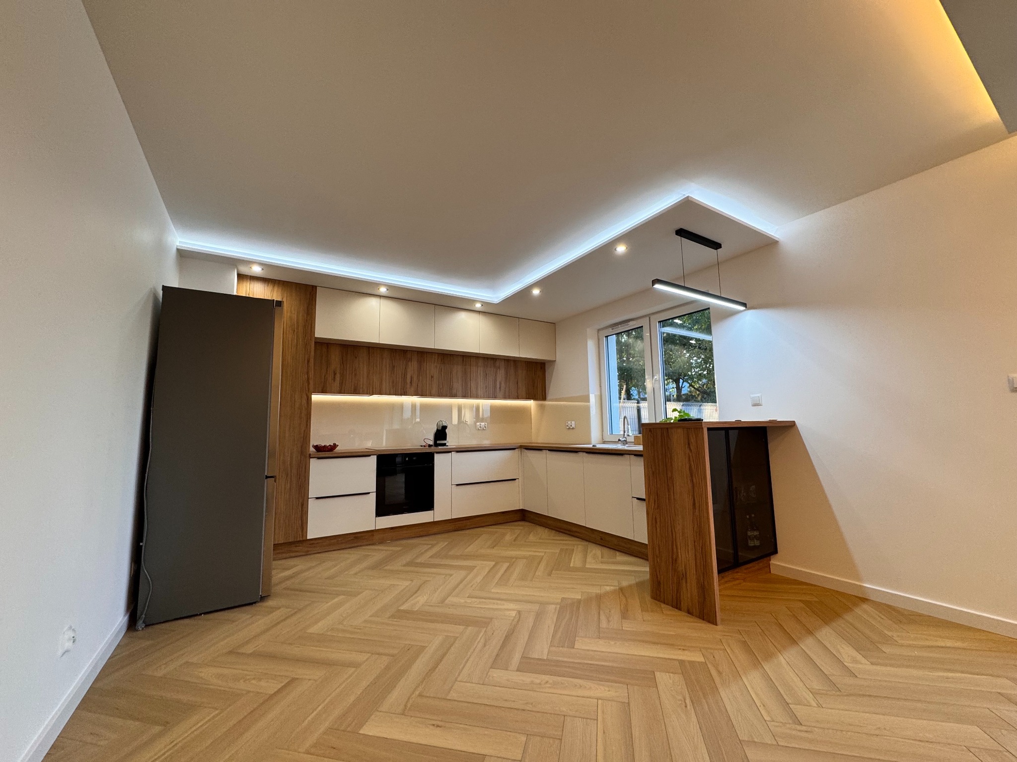 Nowoczesna kuchnia w odcieniach bieli i drewna, parkiet w jodełkę, oświetlenie LED podszafkowe i sufitowe. Minimalistyczny design, widok na zewnątrz przez okno.