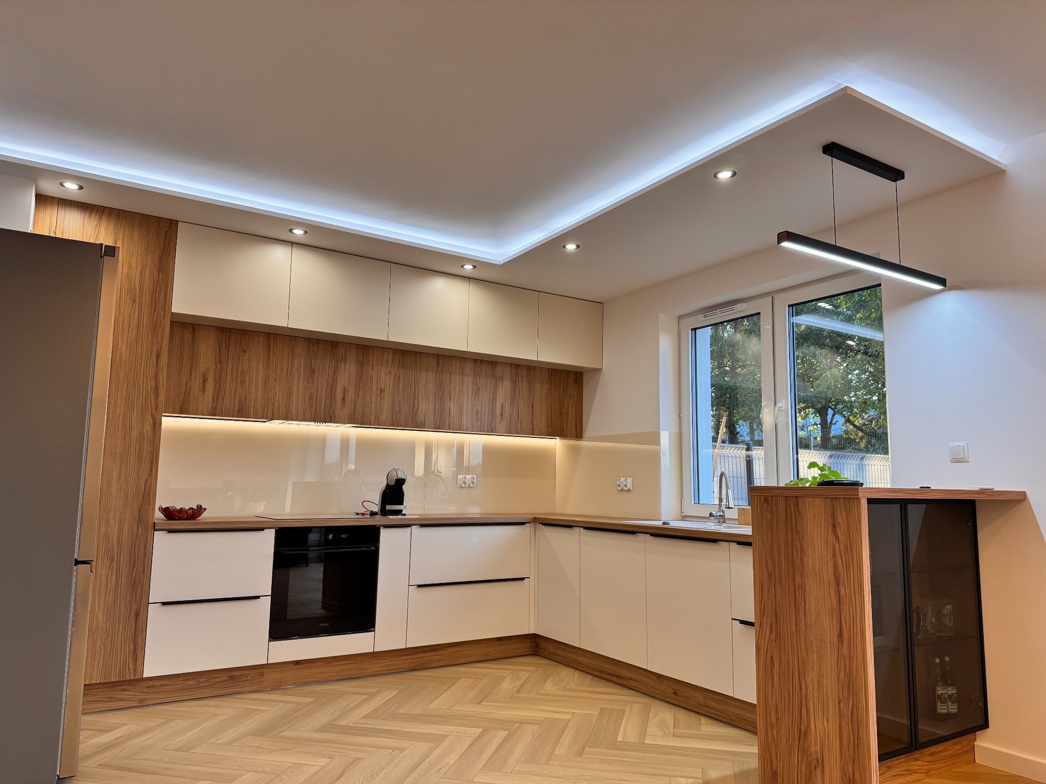 Nowoczesna kuchnia z drewnianymi i białymi szafkami, oświetleniem LED pod sufitem i parkietem w jodełkę. Widok na okno i elementy wyposażenia.