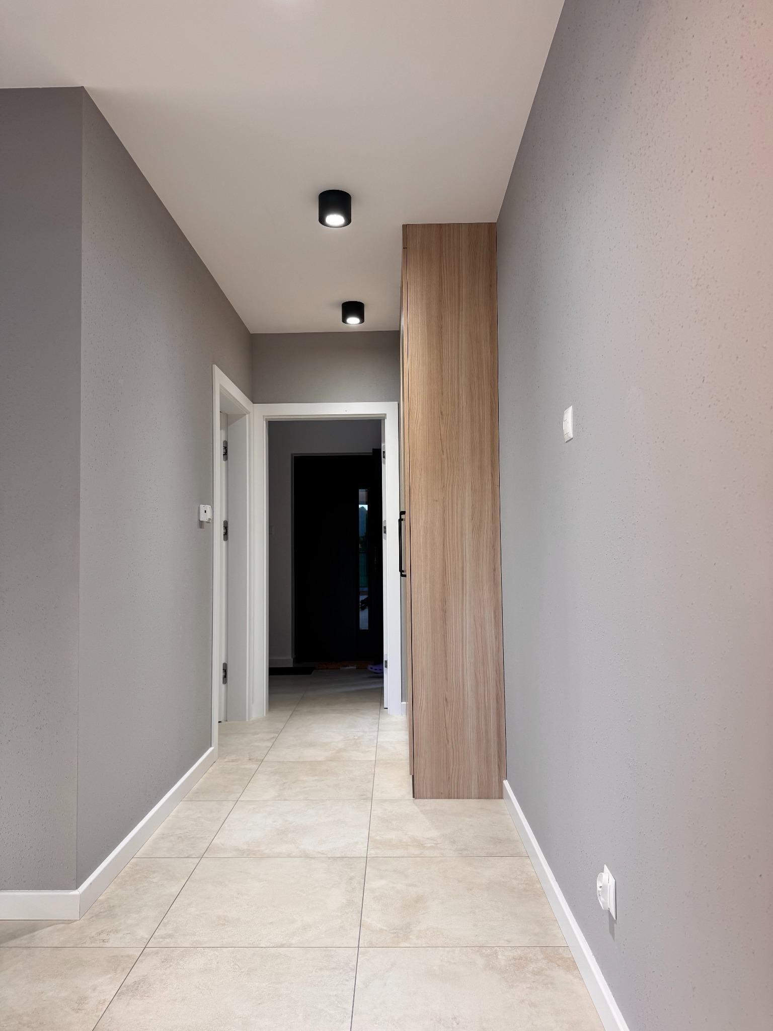 Korytarz po remoncie: szare ściany z fakturą, beżowe płytki podłogowe, białe listwy przypodłogowe, drewniana szafa wnękowa i czarne lampy sufitowe.
