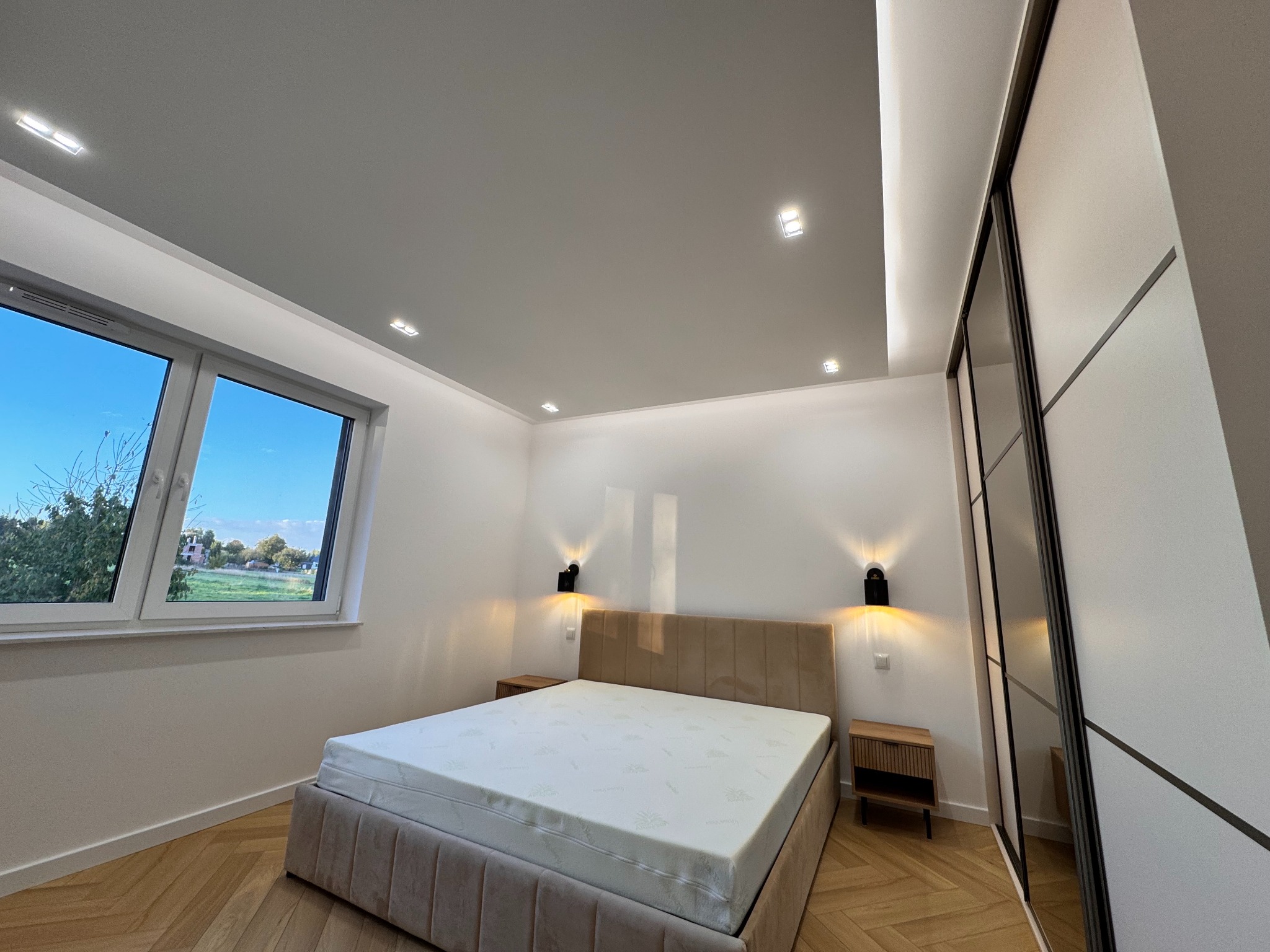 Nowoczesna sypialnia po remoncie: jasne ściany, parkiet w jodełkę, beżowe łóżko z tapicerowanym zagłówkiem, przesuwne drzwi szafy i okno z widokiem na zieleń.