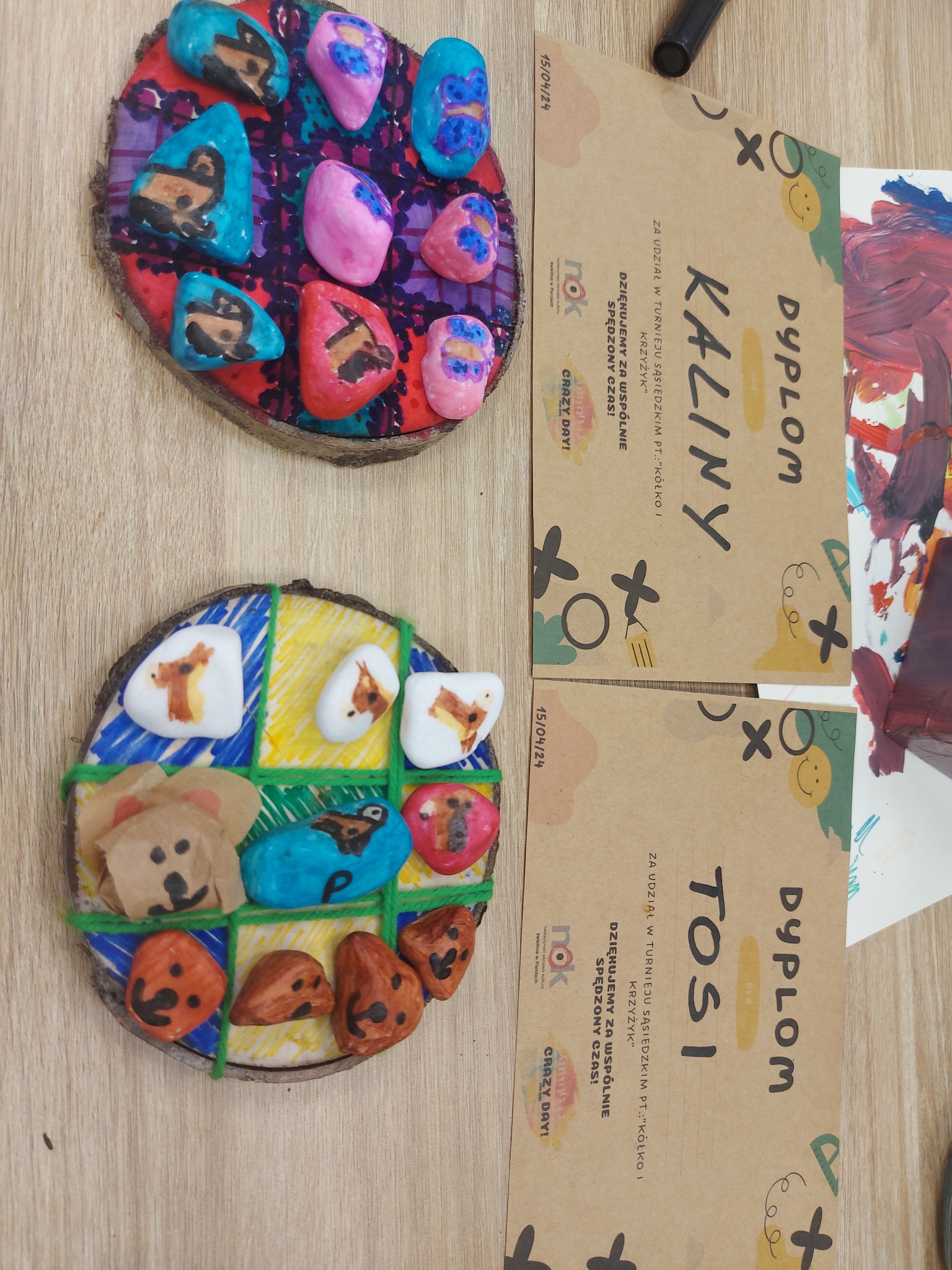 Ręcznie robione dekoracje na plastrach drewna, z pomalowanymi kamieniami przedstawiającymi zwierzęta, obok dyplomy uczestnictwa w warsztatach plastycznych dla dzieci.