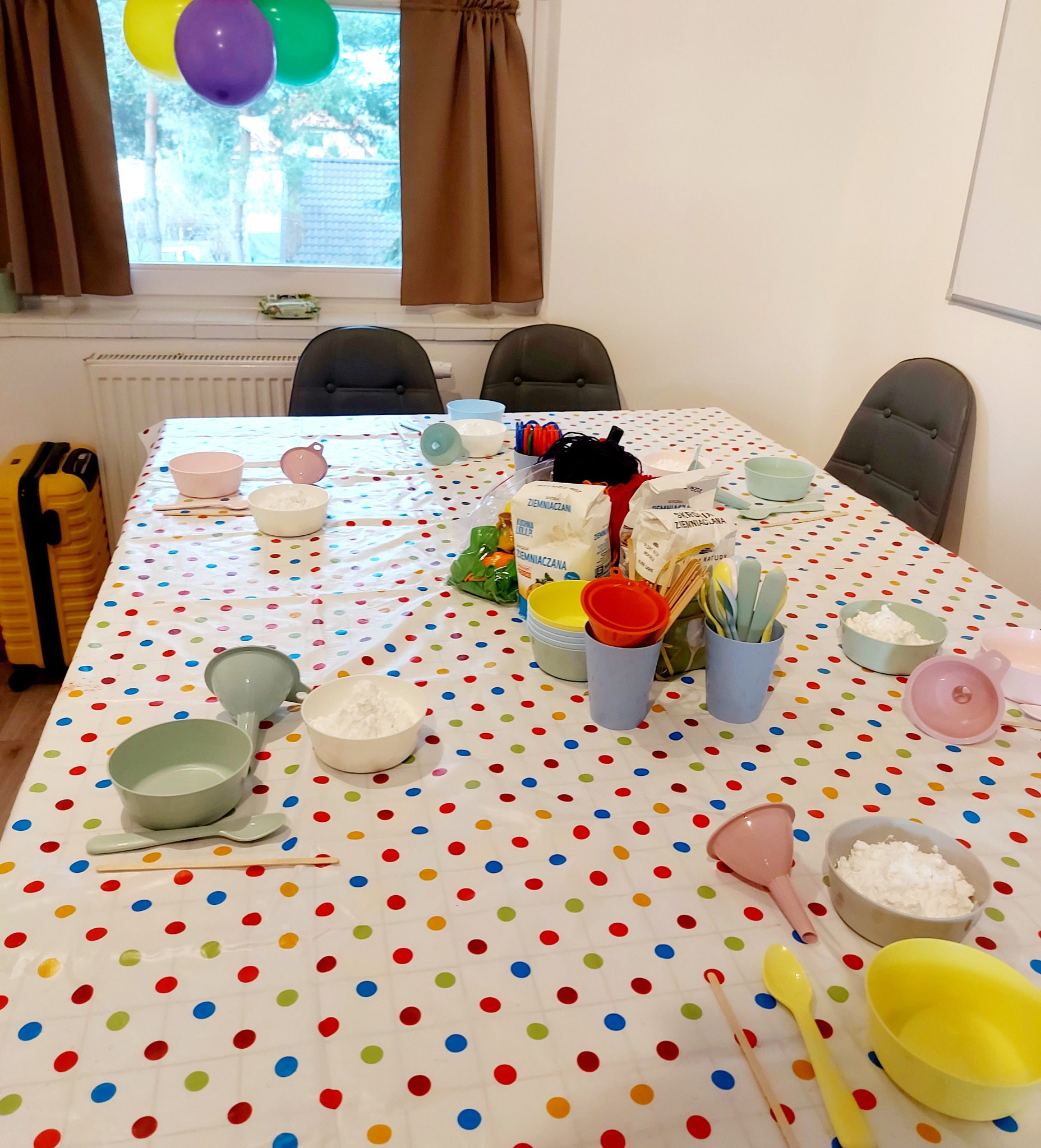 Stół nakryty ceratą w kolorowe kropki, przygotowany do warsztatów kulinarnych dla dzieci, z miseczkami, łyżeczkami, lejkami i opakowaniami mąki ziemniaczanej.