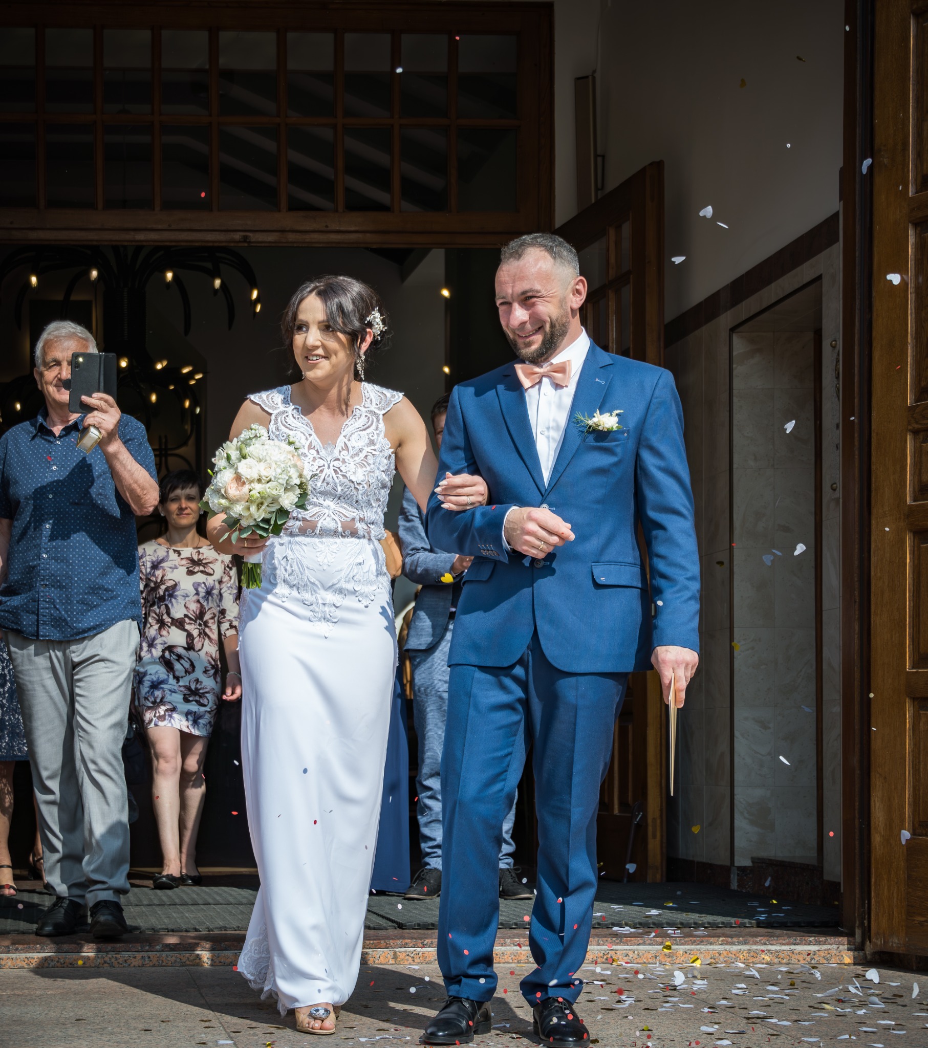 Para młoda opuszcza kościół w strugach konfetti, mężczyzna w niebieskim garniturze z różową muchą, kobieta w białej sukni z koronkową górą i bukietem białych kwiatów.