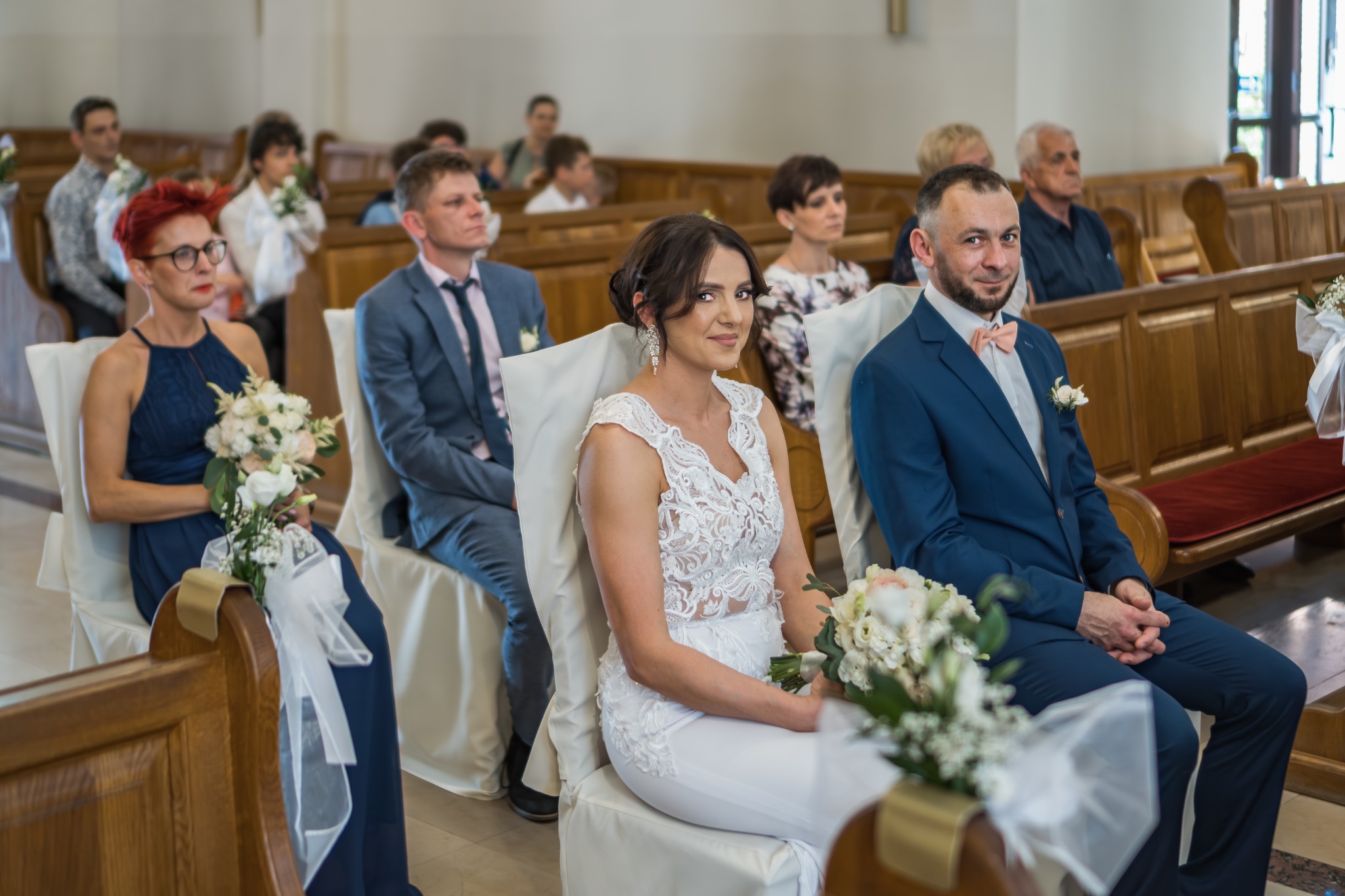 Para młoda siedzi w kościele podczas ceremonii ślubnej, panna młoda w koronkowej sukni, pan młody w niebieskim garniturze i muszce, w tle goście i drewniane ławki.