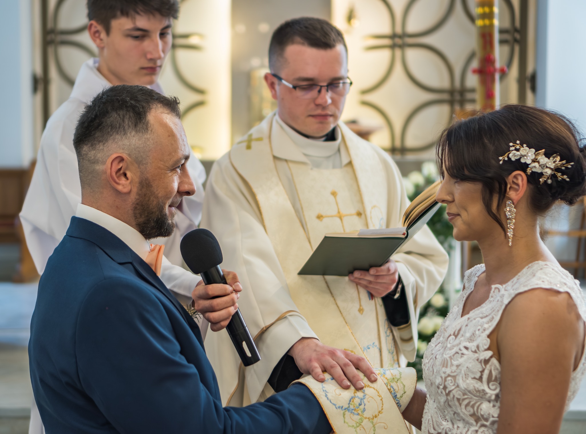 Para młoda podczas ceremonii ślubnej w kościele, pan młody trzyma mikrofon, a ksiądz czyta z księgi.