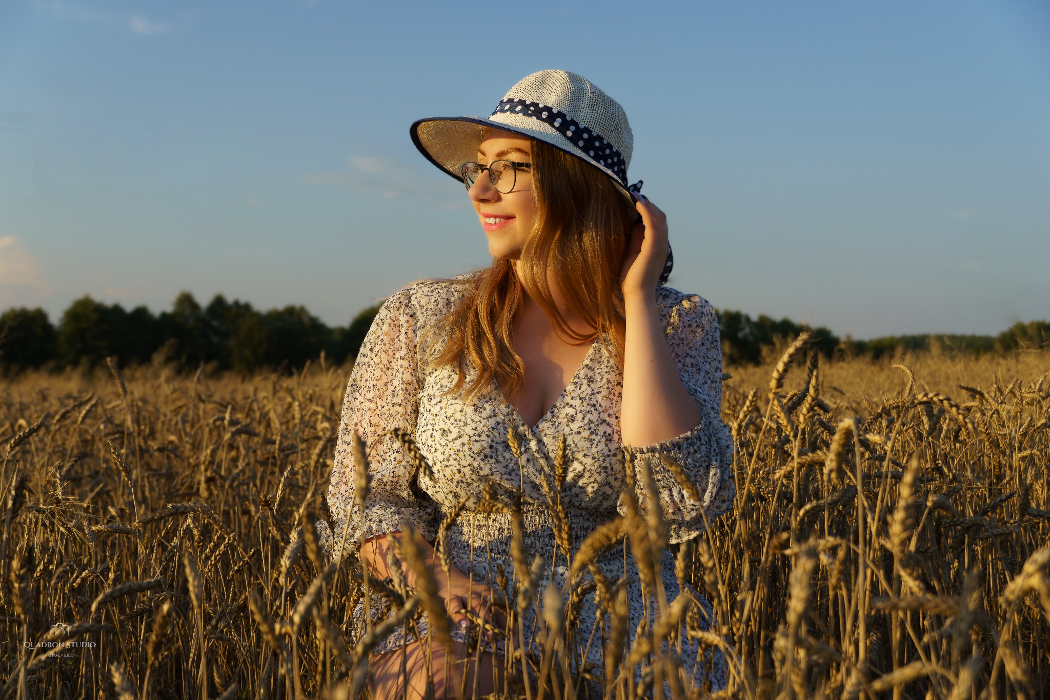 Portret kobiety w sukience w kwiaty i kapeluszu stojącej pośród dojrzewającego pola zboża w ciepłym świetle zachodzącego słońca.