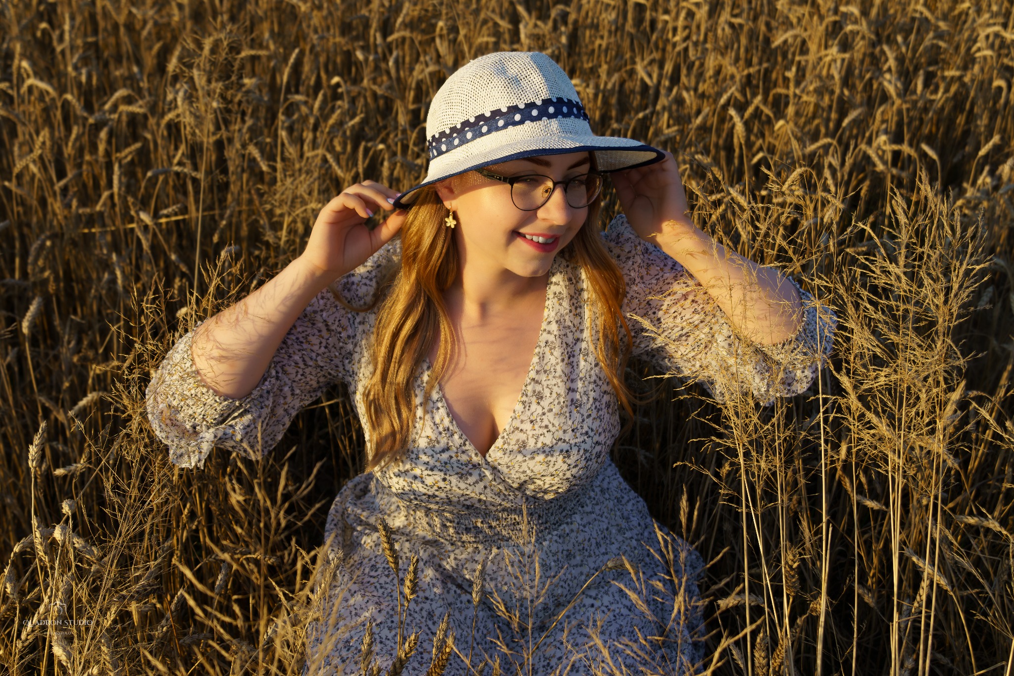 Uśmiechnięta blondynka w okularach i kapeluszu z niebieską wstążką, leżąca w polu zboża w sukience w kwiaty, delikatne złote światło.