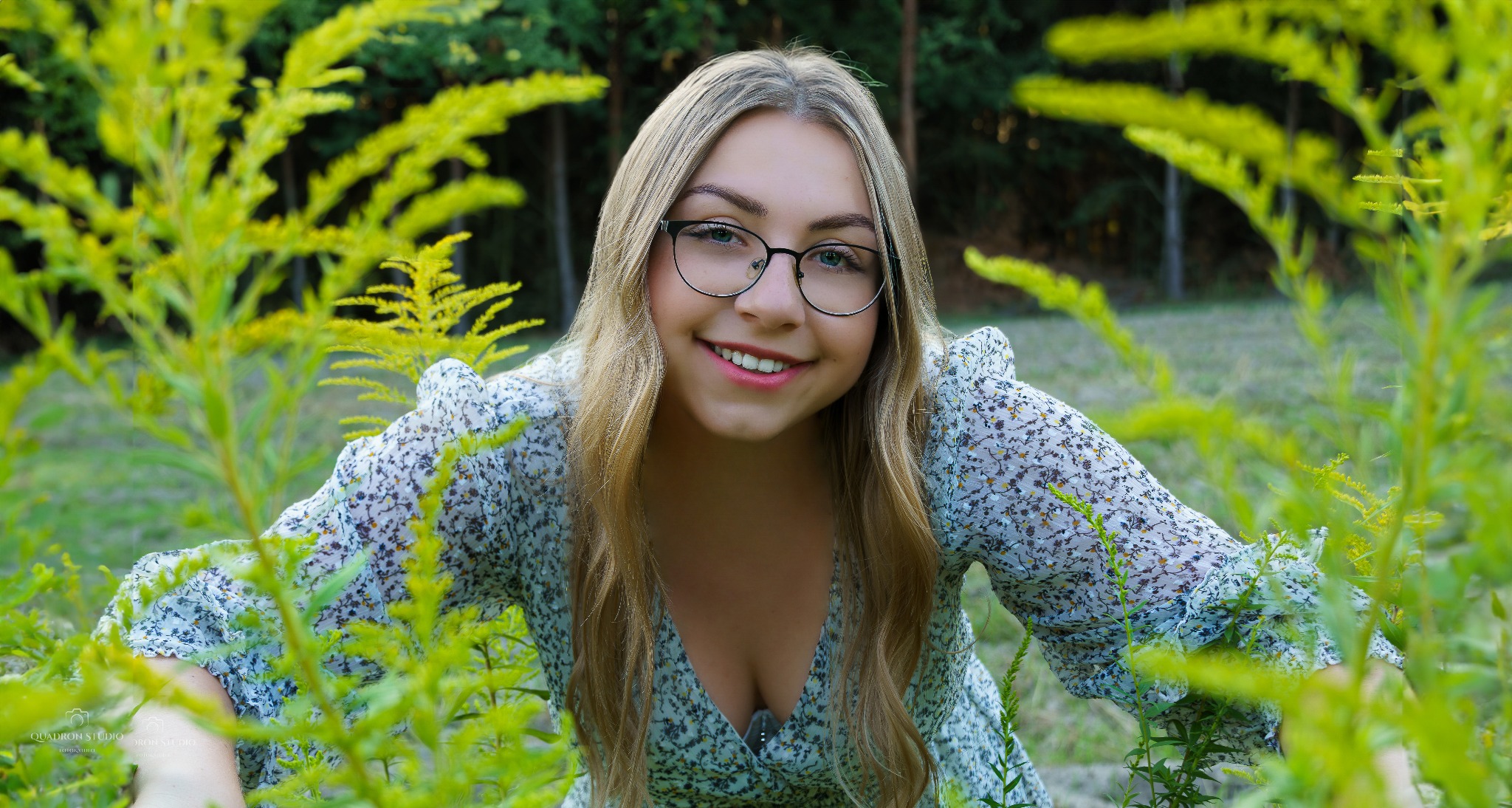 Portret uśmiechniętej młodej kobiety w okularach, w kwiecistej sukience, pozującej w plenerze wśród zielonej roślinności.
