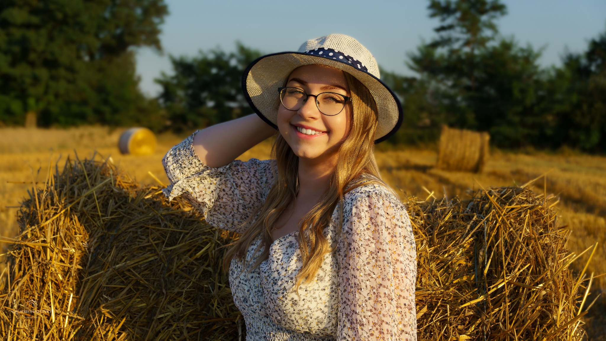 Uśmiechnięta blondynka w okularach i kapeluszu, oparta o snop siana na polu w słoneczny dzień.