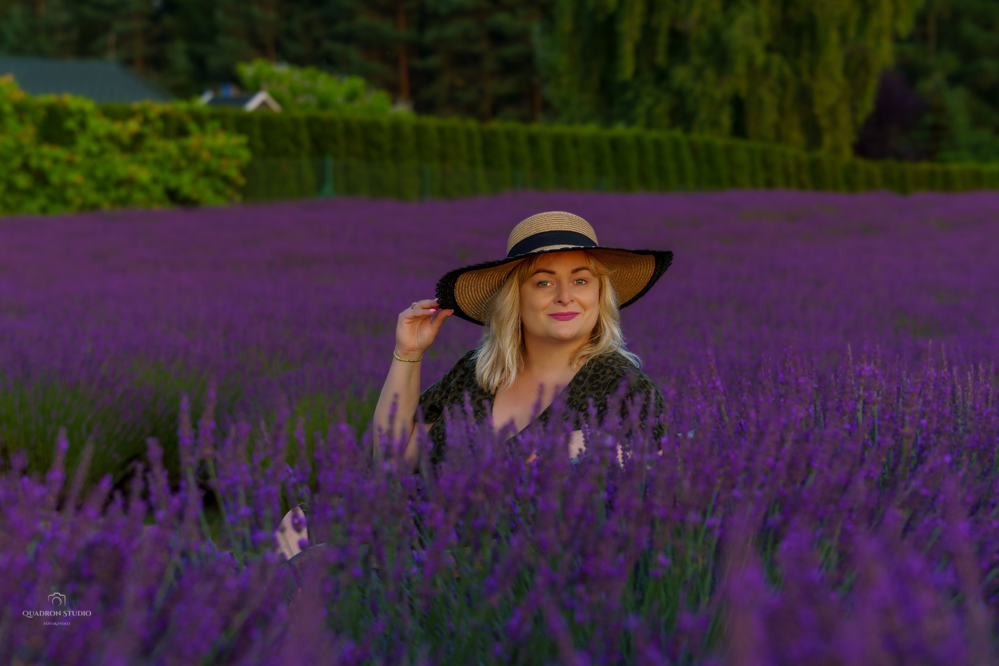 Uśmiechnięta blondynka w słomkowym kapeluszu z czarnym rondem pozuje na polu lawendy, trzymając rondo kapelusza, w tle zielone krzewy i dom.