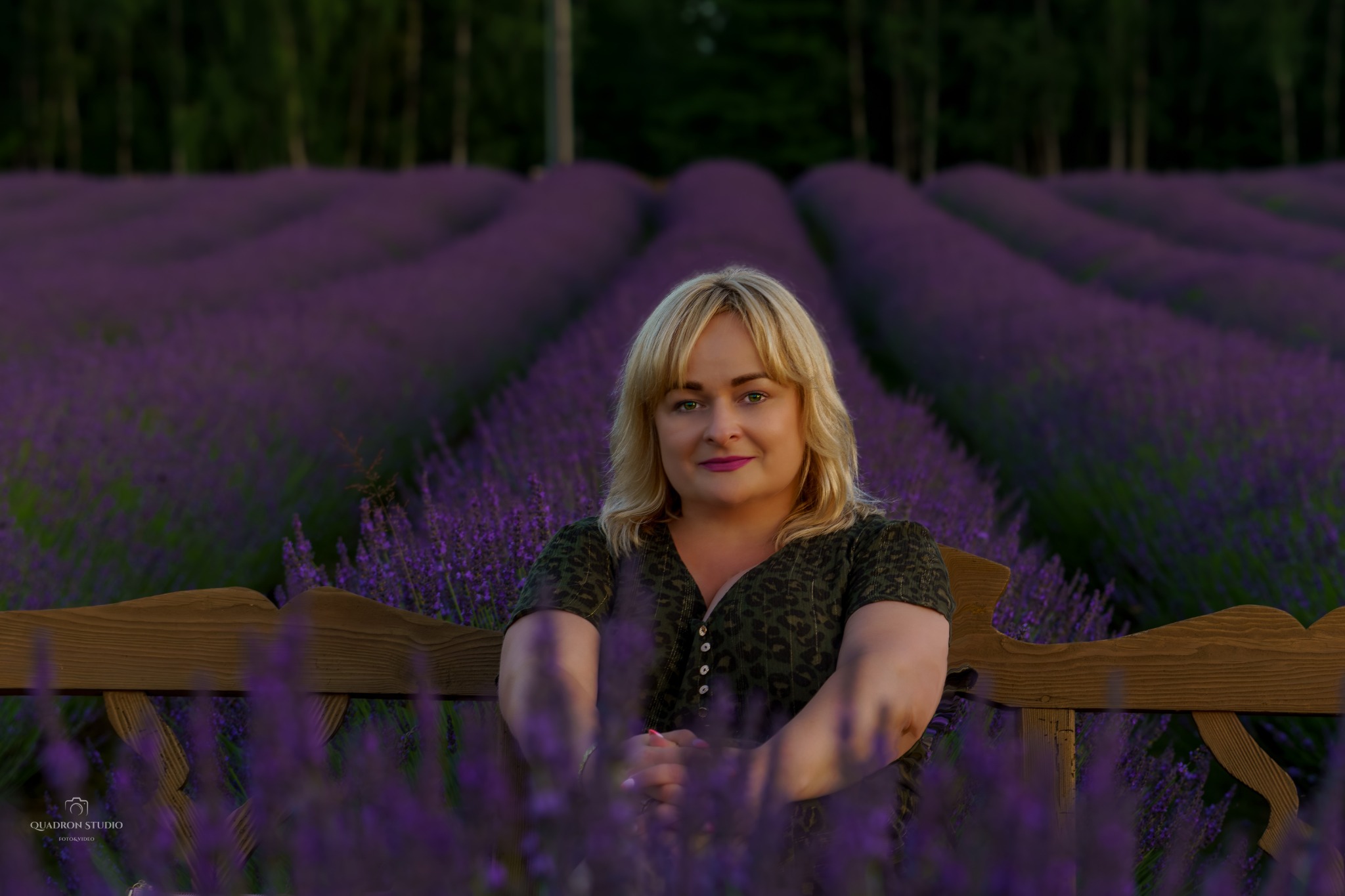 Kobieta z blond włosami siedzi na drewnianej ławce na tle pola kwitnącej lawendy, z delikatnym uśmiechem i spojrzeniem w obiektyw.