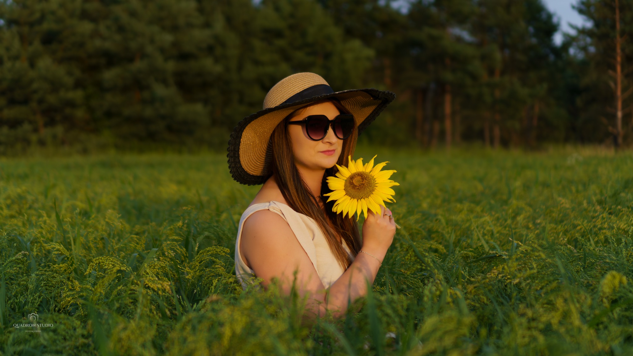 Kobieta w słomkowym kapeluszu i okularach przeciwsłonecznych trzymająca słonecznik na tle pola i lasu.