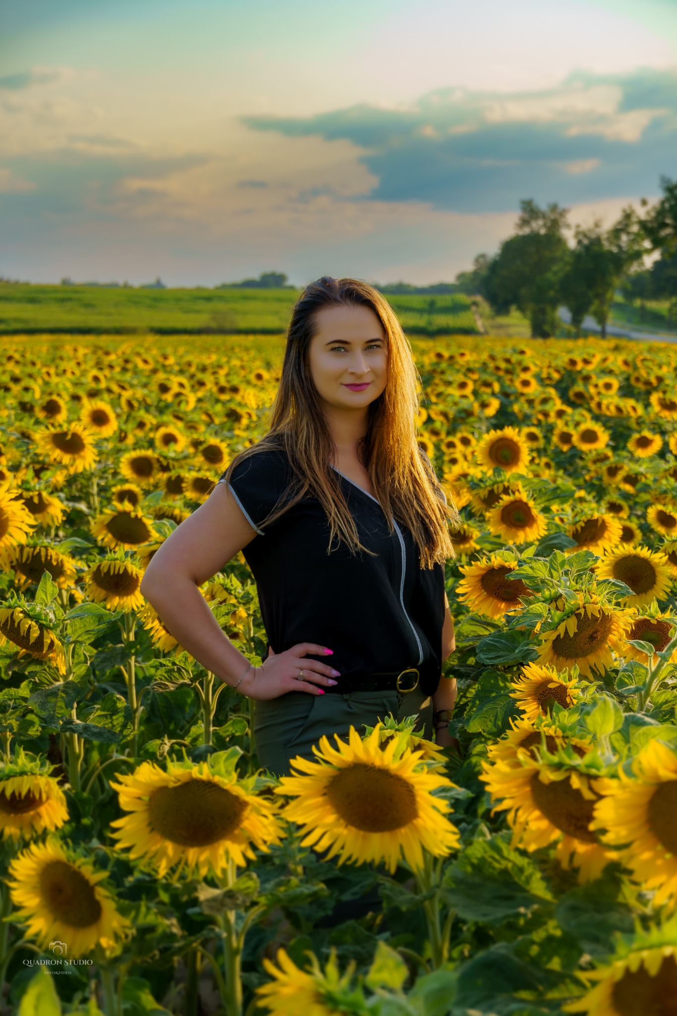 Portret kobiety w czarnej bluzce i zielonych spodniach stojącej na polu słoneczników w promieniach zachodzącego słońca, z widocznym horyzontem i drzewami w tle.