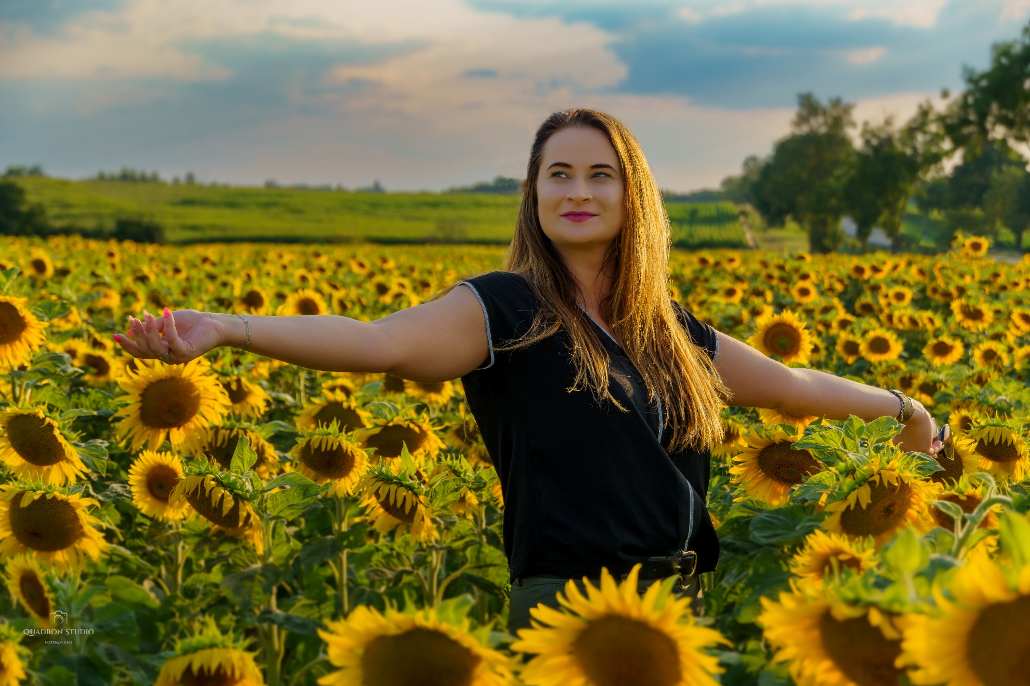 Kobieta z rozłożonymi ramionami stoi na polu słoneczników w ciepłym, popołudniowym świetle, z widocznymi drzewami i polami w oddali.
