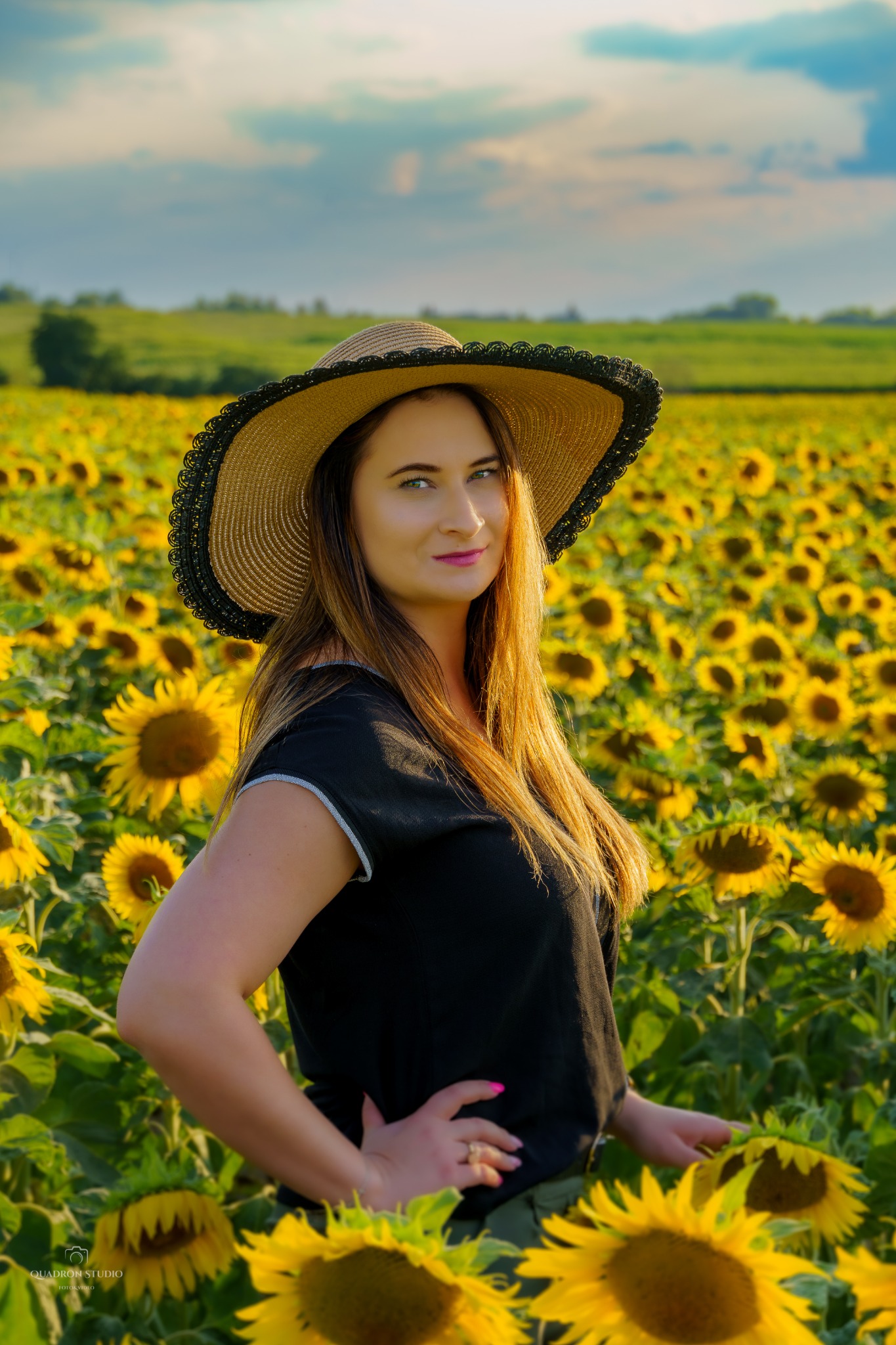 Portret kobiety w słomkowym kapeluszu z czarną koronką, stojącej na polu słoneczników w promieniach zachodzącego słońca.
