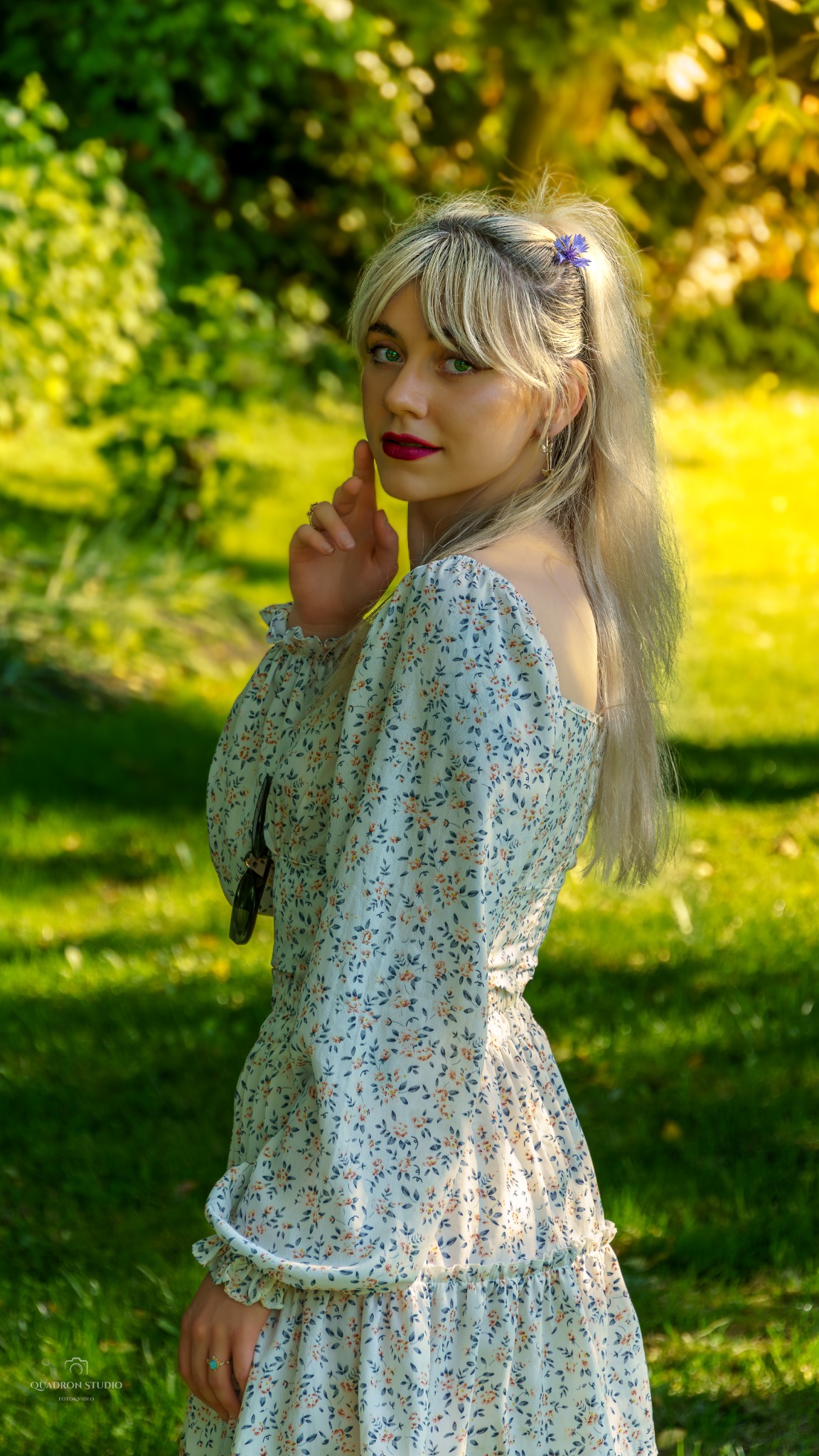 Portret kobiety z blond grzywką i kwiatem we włosach, w kwiecistej sukience na tle zieleni parku, spoglądającej przez ramię.