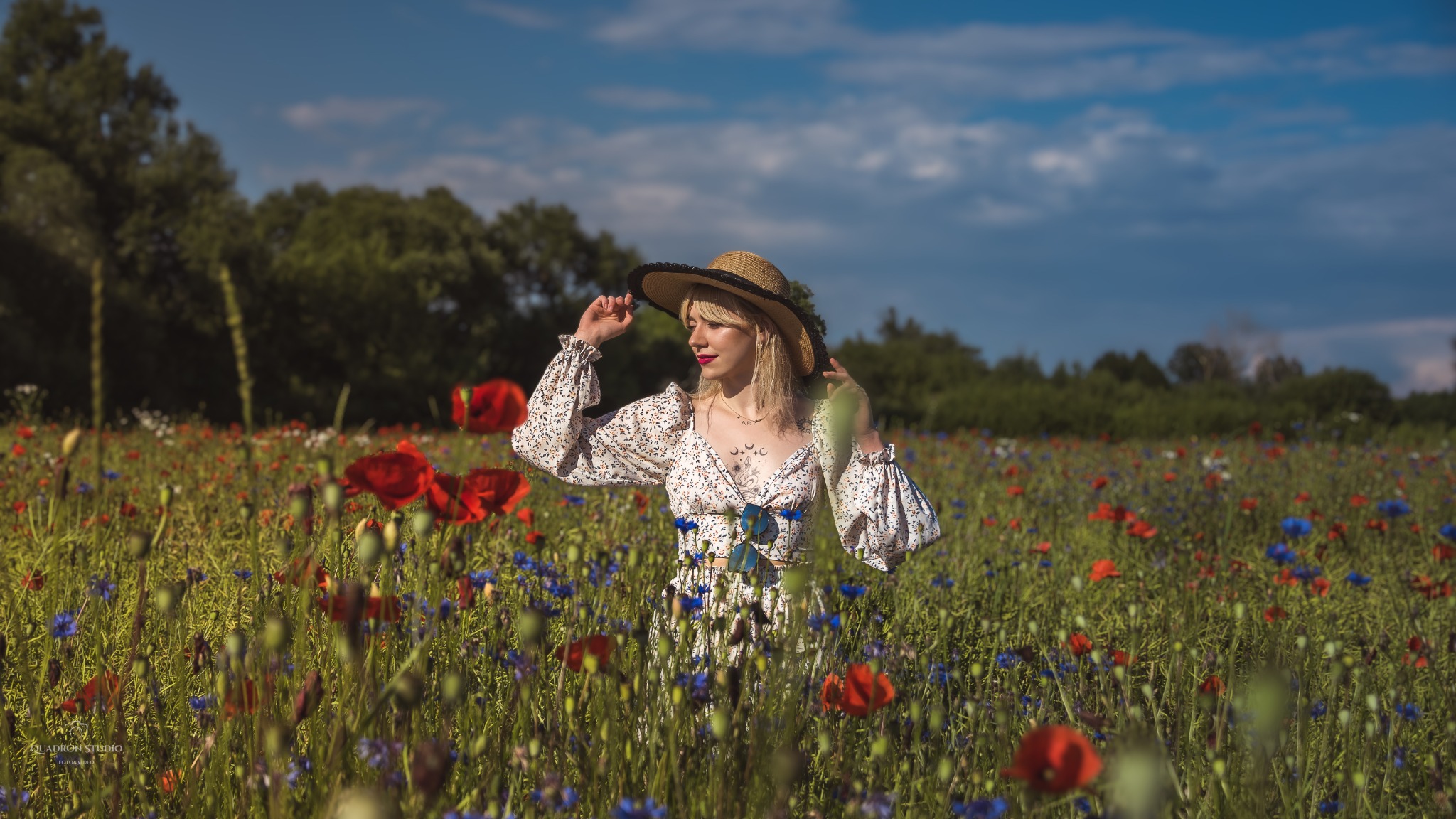 Blondwłosa kobieta w kwiecistej sukience i kapeluszu stoi na łące pełnej maków i chabrów, delikatnie trzymając rondo kapelusza.