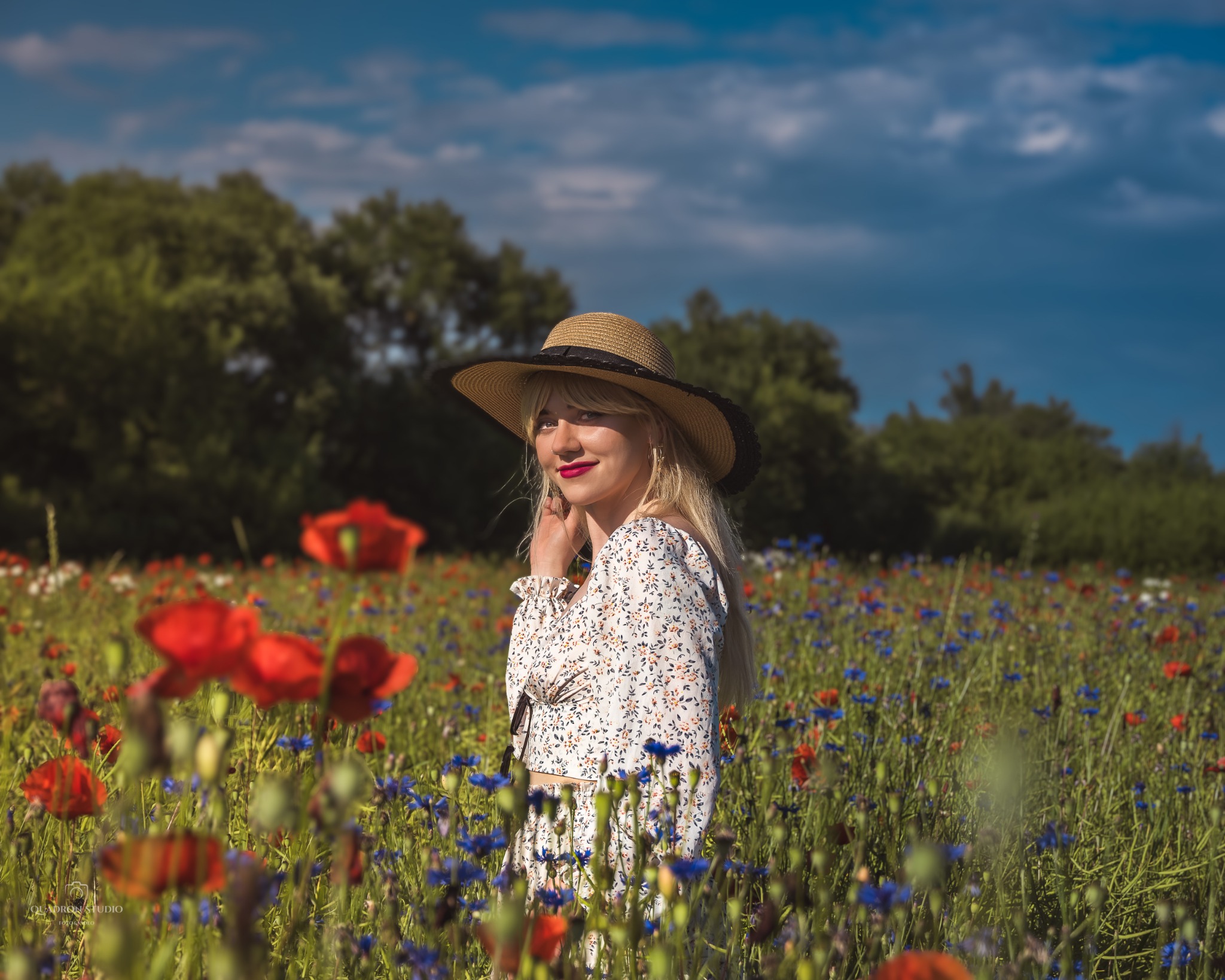 Portret kobiety w kwiecistej sukience i słomkowym kapeluszu na łące z makami i chabrami, z drzewami i błękitnym niebem w tle.