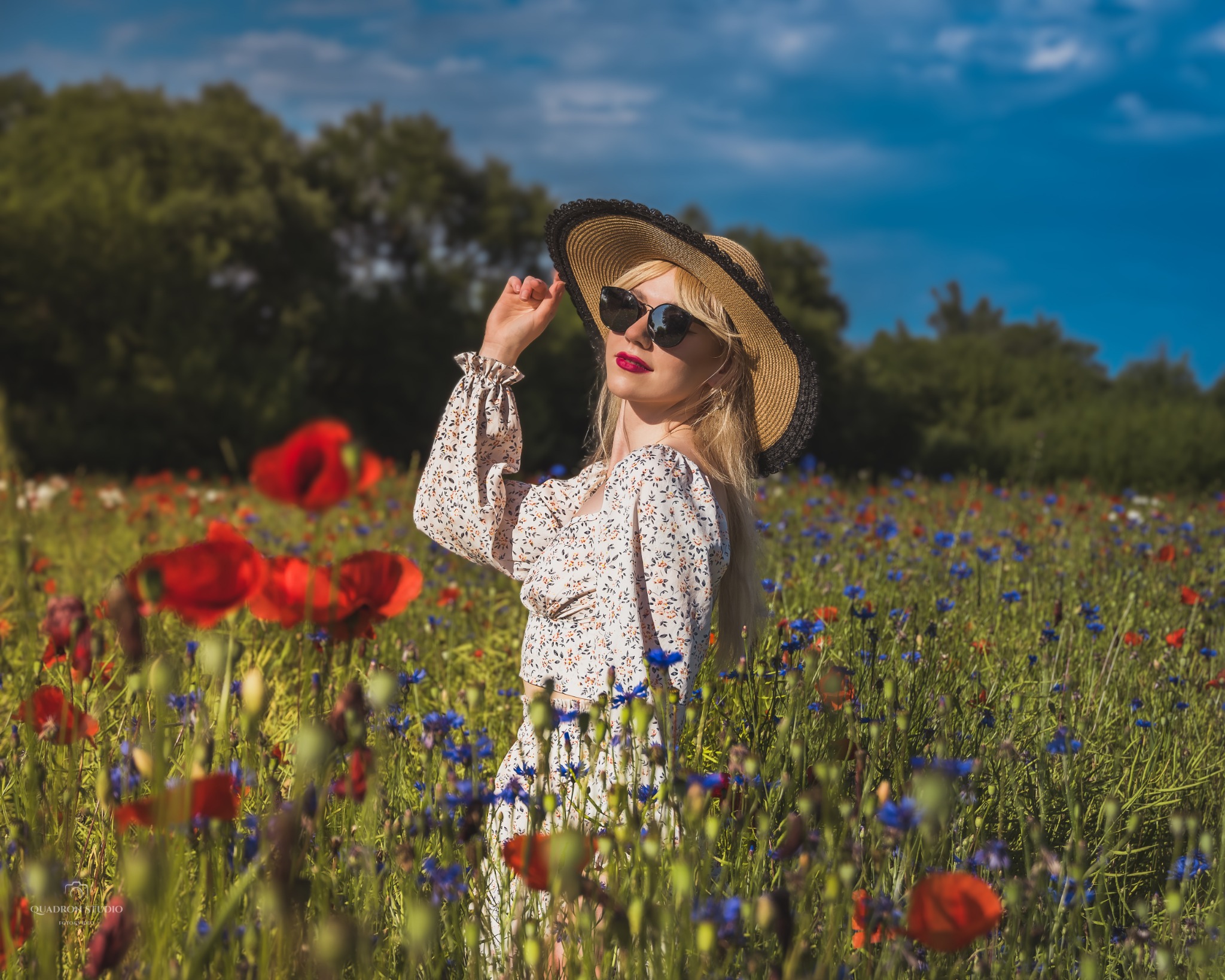 Blondwłosa kobieta w kwiecistym stroju i słomkowym kapeluszu z czarną obwódką, pozuje na łące pełnej maków i chabrów, w słoneczny dzień.
