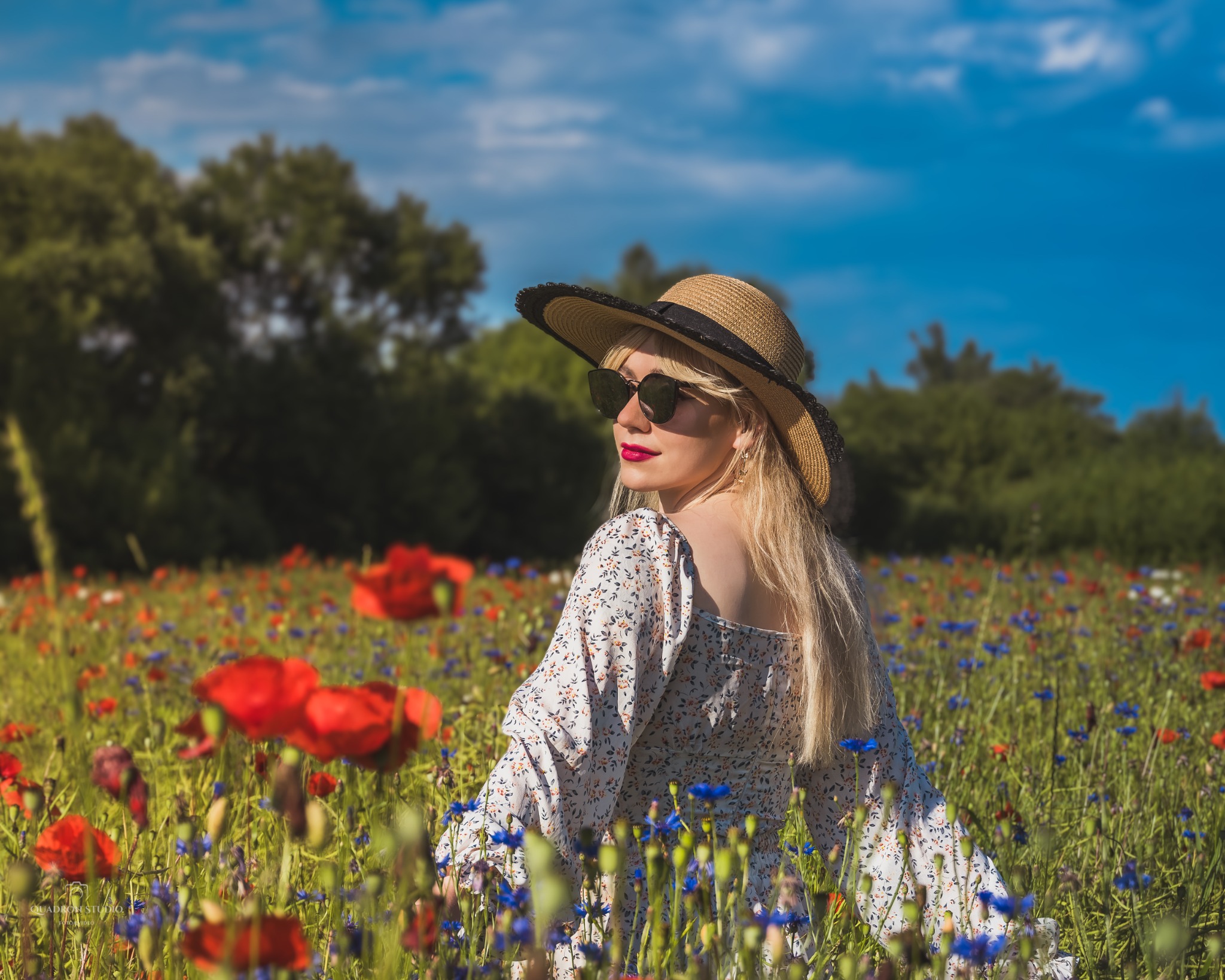 Kobieta w kapeluszu i okularach przeciwsłonecznych, odwrócona tyłem, pozuje na łące z makami i chabrami.