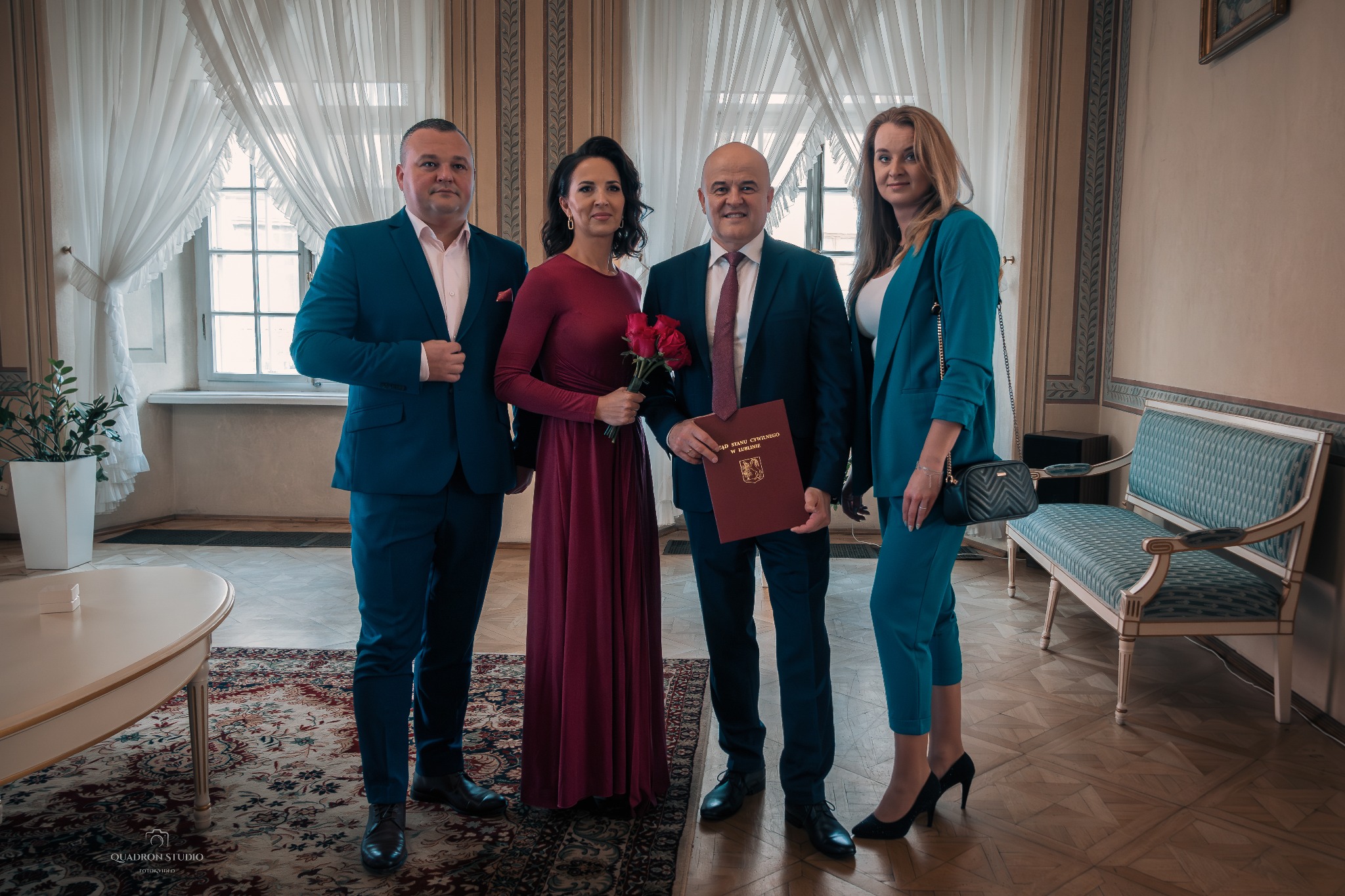 Grupa elegancko ubranych osób, w tym kobieta z bukietem czerwonych róż i mężczyzna trzymający akt ślubu, pozuje na tle jasnego wnętrza z zasłonami i wzorzystym dywanem.
