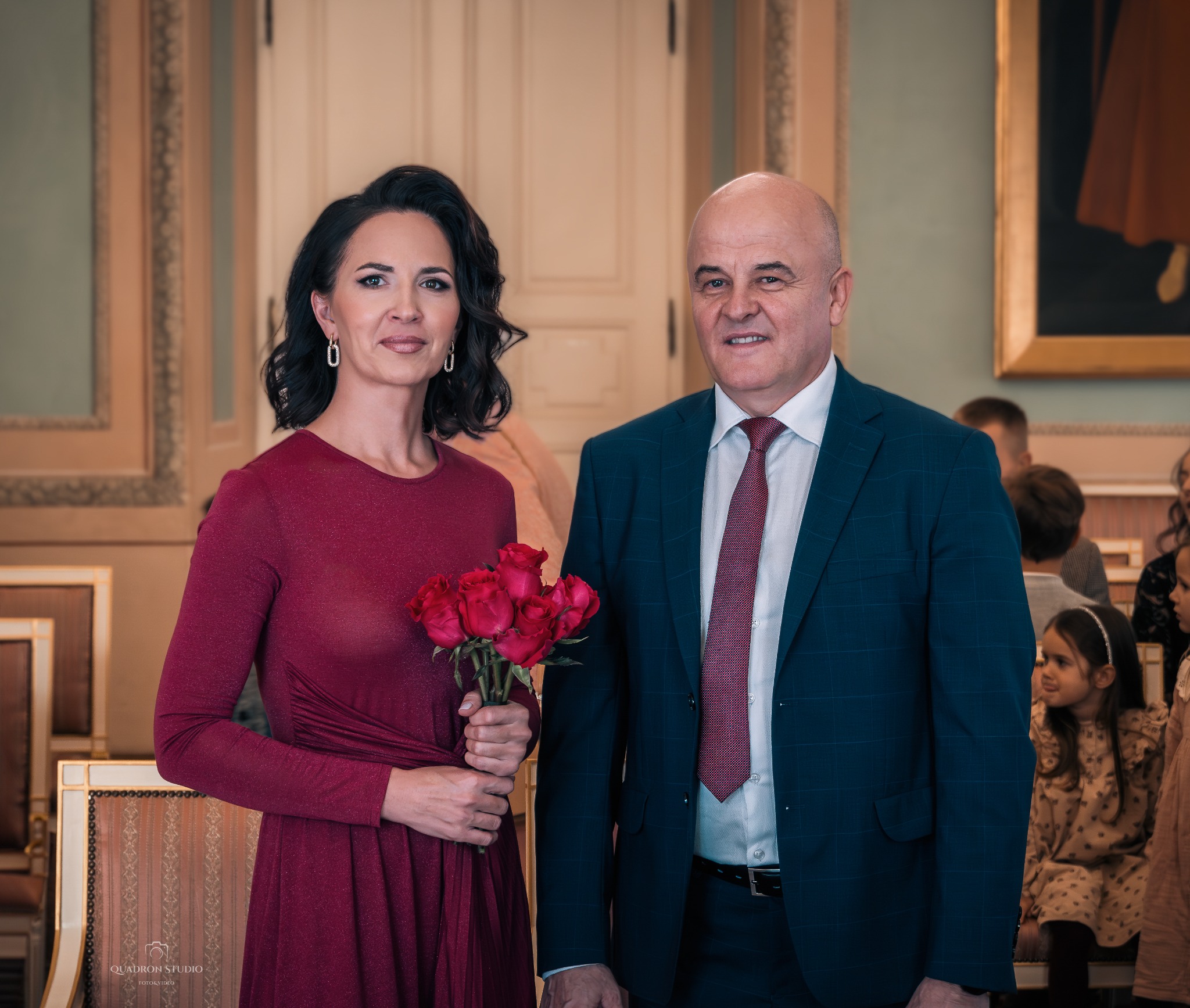Elegancka para: kobieta w bordowej sukience trzyma bukiet czerwonych róż, mężczyzna w granatowym garniturze z bordowym krawatem, w tle dzieci i dekoracyjne wnętrze.