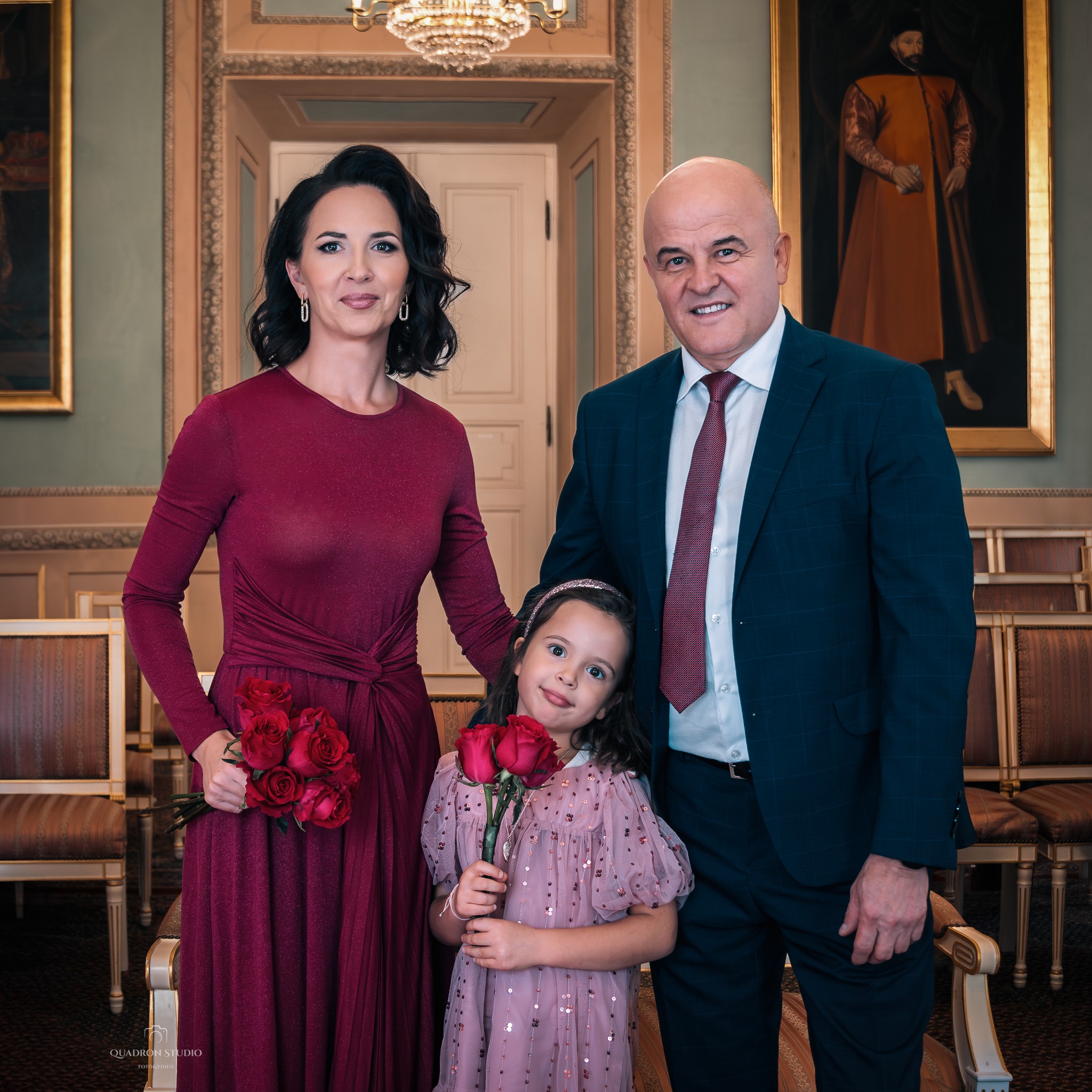 Elegancka, uśmiechnięta rodzina: matka w bordowej sukni z bukietem czerwonych róż, ojciec w granatowym garniturze i córka w różowej sukience z brokatem, trzymająca dwie róże, pozują w bogato...