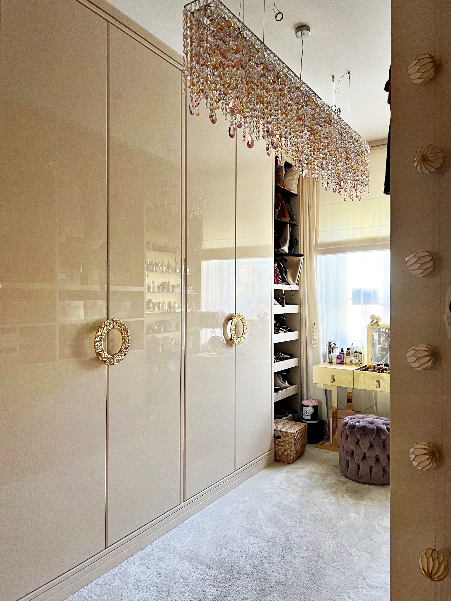 Beżowa, lakierowana szafa wnękowa z okrągłymi, zdobionymi uchwytami, oświetlona kryształowym żyrandolem w garderobie z toaletką.