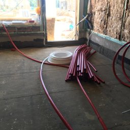 Sh&K - Wnętrze w trakcie prac instalacyjnych: wiązka czerwonych rur grzewczych na podłodze, biała zwinięta rura obok, ocieplone wełną mineralną ściany, widok na plac budowy przez okno.