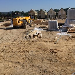 Sh&K - Budowa domów jednorodzinnych w stanie surowym otwartym, widoczny żółty wózek widłowy JCB, materiały budowlane i pracownicy w oddali.