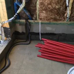 Ściana w trakcie prac instalacyjnych: widoczne przewody elektryczne w peszlach, izolacja z wełny mineralnej, oraz stos czerwonych rur karbowanych na podłodze.