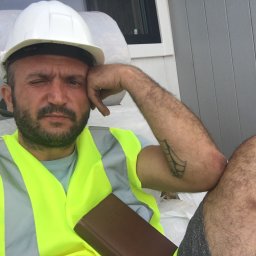 Sh&K - Pracownik budowlany w kasku i kamizelce odblaskowej, z przymrużonym okiem, oparty o rękę, z kalendarzem w dłoni, na tle folii i szarej ściany.