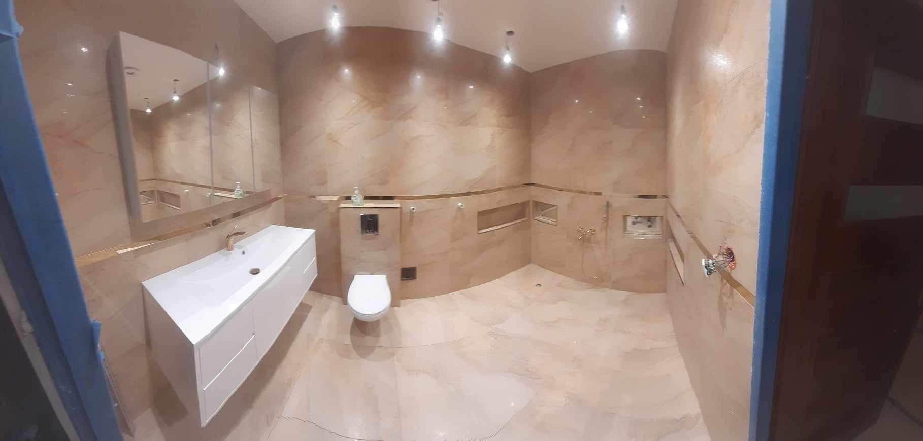 Wykończona łazienka z beżowymi marmurkowymi płytkami, podwieszana biała umywalka z szafką, wisząca muszla klozetowa, oświetlenie punktowe i lustro z podświetleniem.