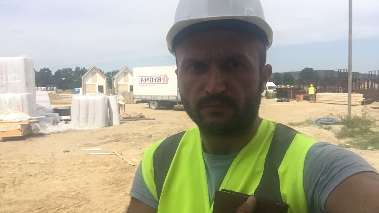 Autoportret pracownika budowlanego w kasku i kamizelce odblaskowej na placu budowy z widocznymi materiałami izolacyjnymi, elementami konstrukcji domów szkieletowych i transportem w tle.