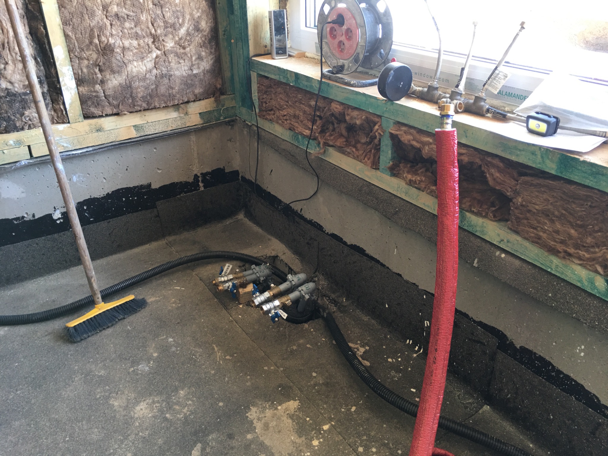 Przygotowanie do wykończenia podłogi w pomieszczeniu: izolacja ścian wełną mineralną, instalacja rur z zaworami oraz czarna izolacja przeciwwilgociowa na podłodze, obok stoi miotła.