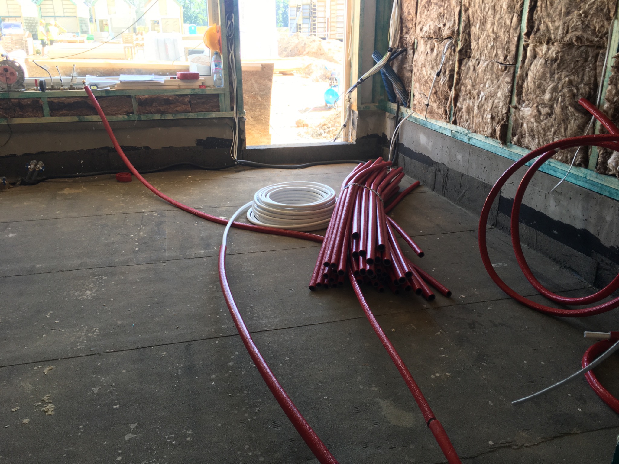 Wnętrze w trakcie prac instalacyjnych: wiązka czerwonych rur grzewczych na podłodze, biała zwinięta rura obok, ocieplone wełną mineralną ściany, widok na plac budowy przez okno.